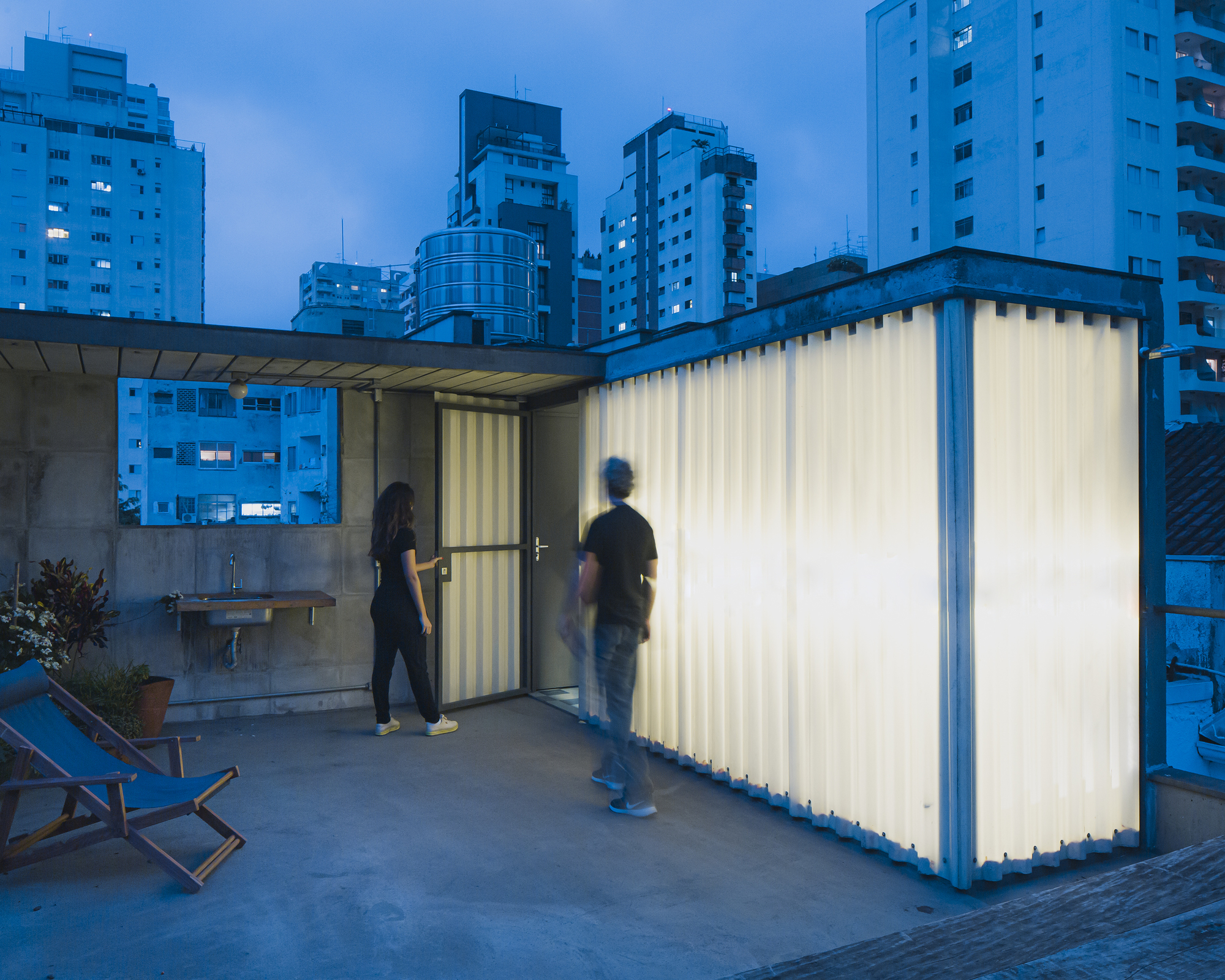 Rooftop terrace with backlit corrugated panel enclosure and two figures at dusk