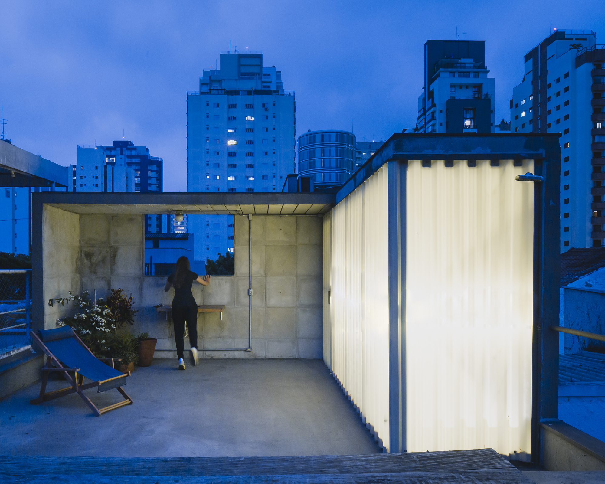 Rooftop terrace with translucent corrugated walls and glass pavilion at dusk with city skyline beyond