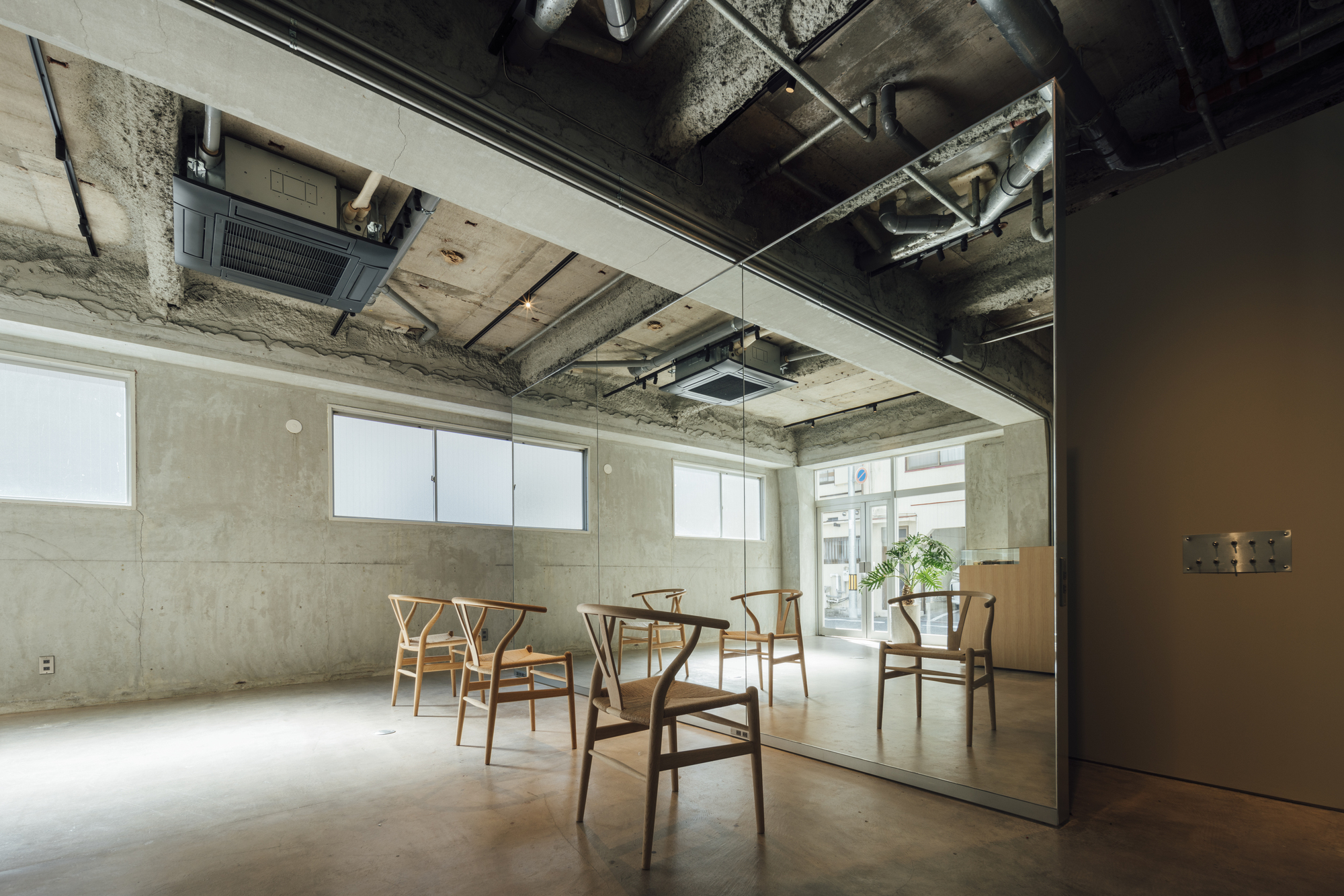 Glass-enclosed room with exposed concrete walls and ceiling ducts reflecting wooden chairs on polished floor
