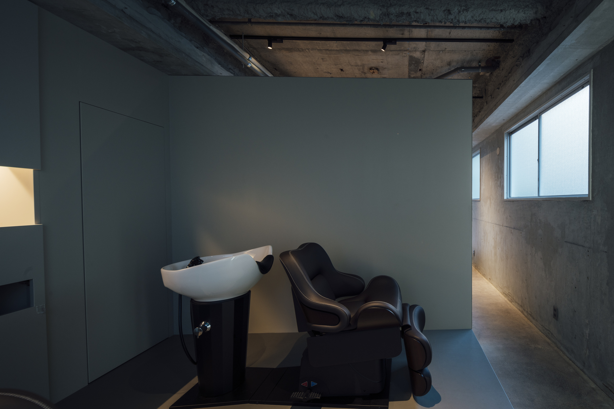 Salon space with wall-mounted washbasin, reclining chair, and concrete surfaces lit by natural light