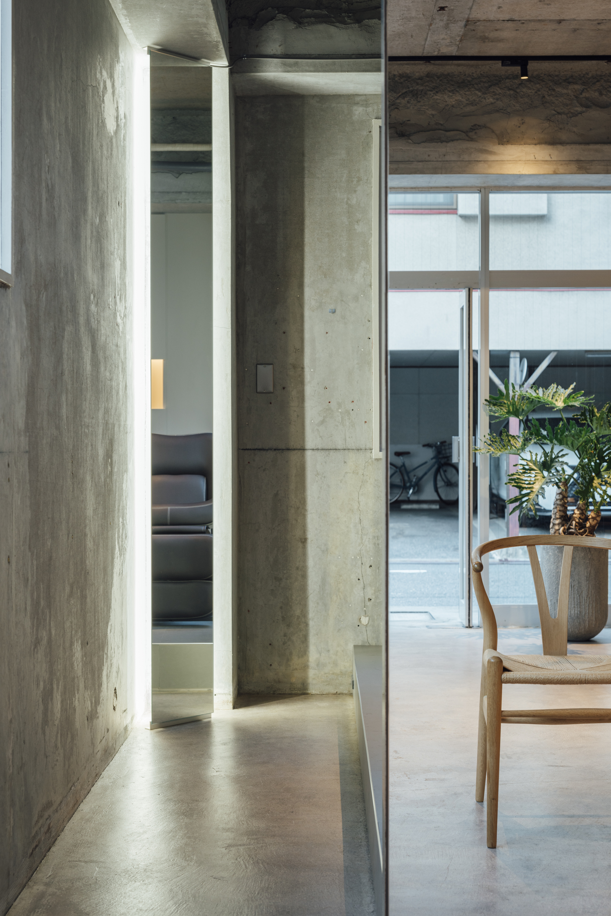 Corridor with exposed concrete walls and ceiling leading past a wooden chair to a glazed courtyard