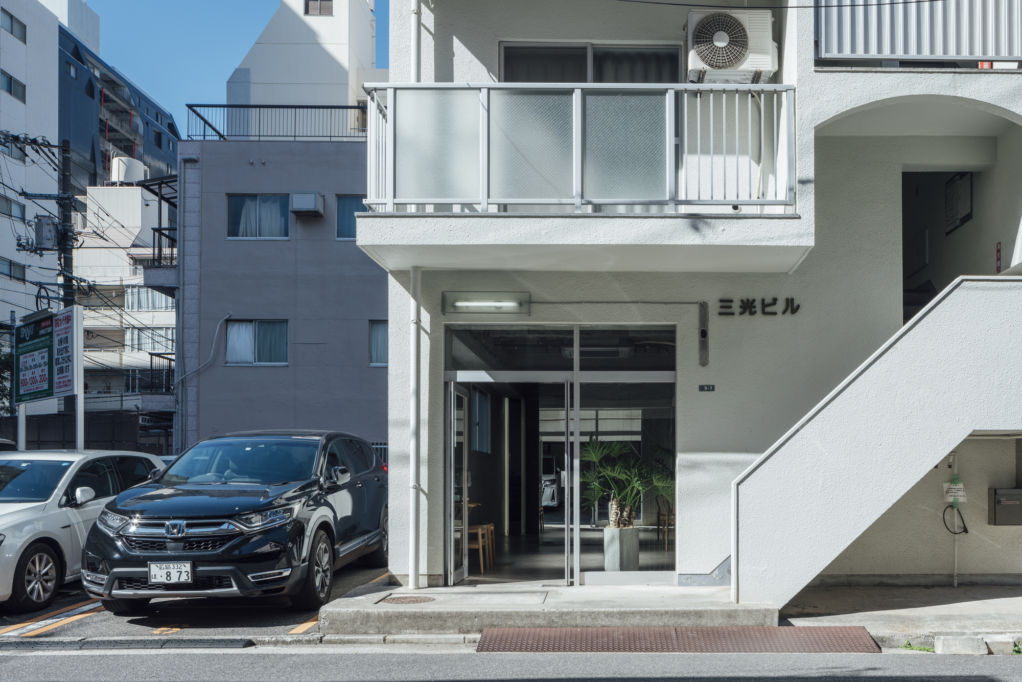 Street facade with glass entry doors and metal balcony railing above parked cars in daylight