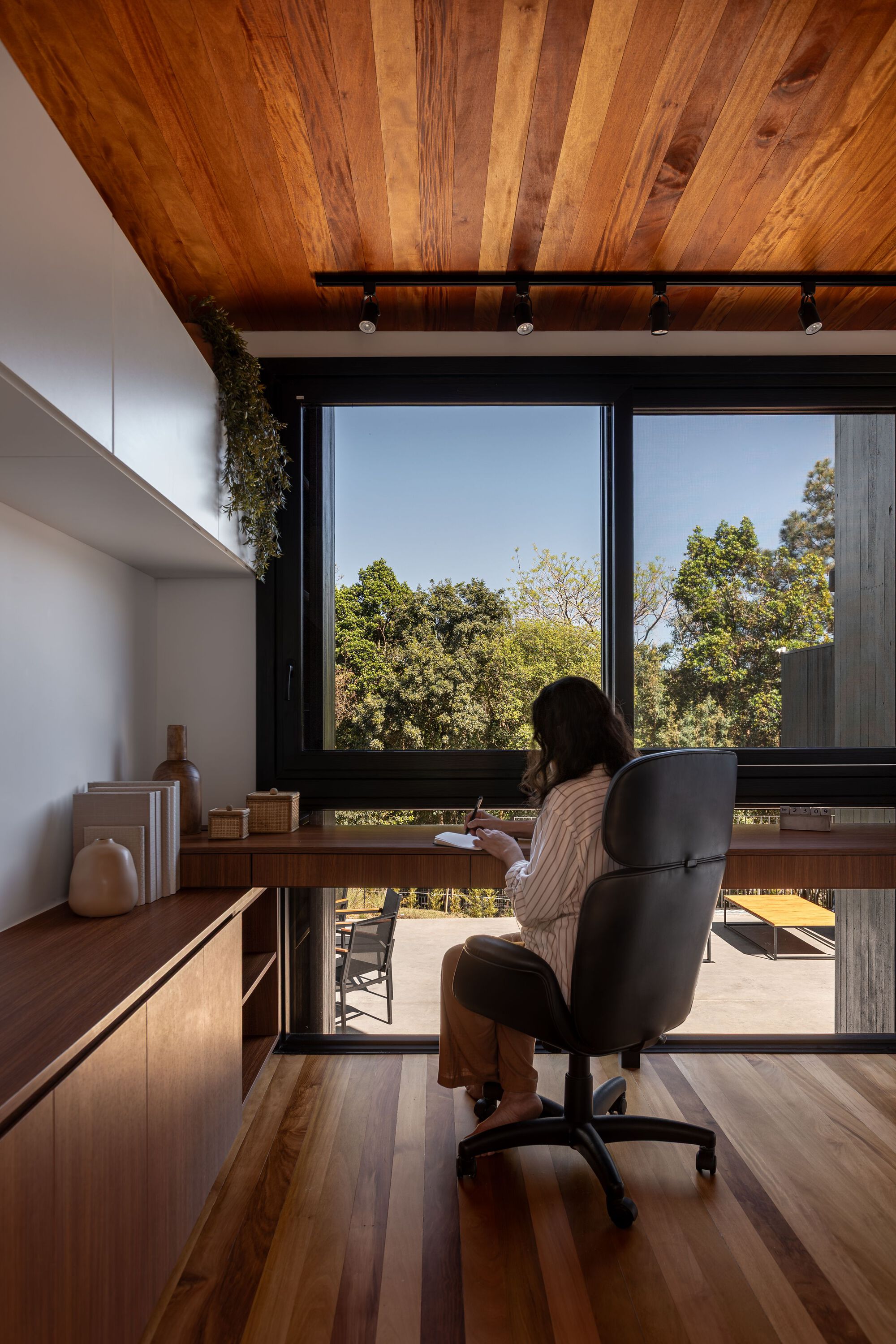 Home office with timber desk facing floor-to-ceiling windows overlooking green treetops in daylight