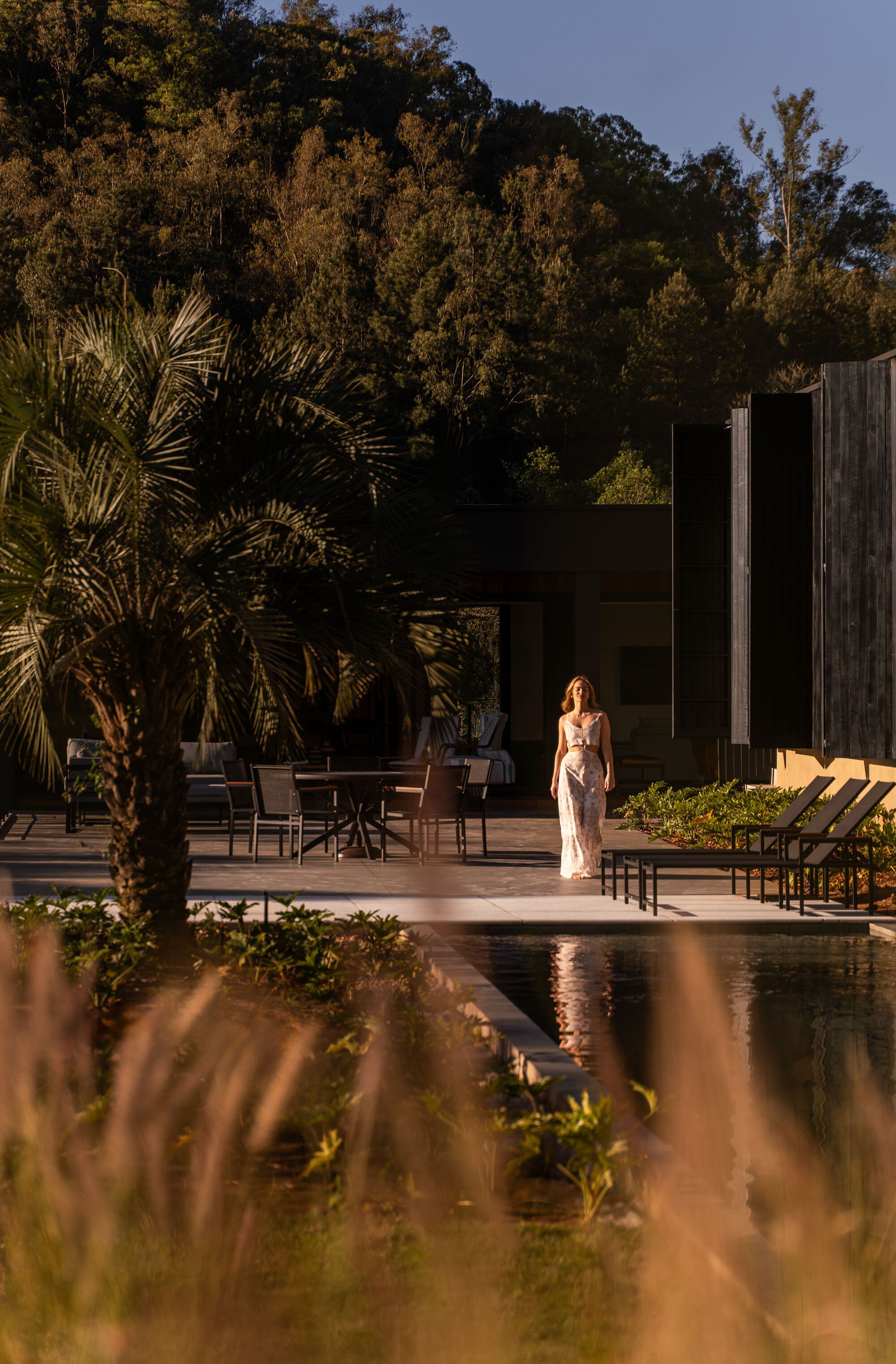 Poolside terrace at dusk with a figure standing near outdoor furniture and a palm tree