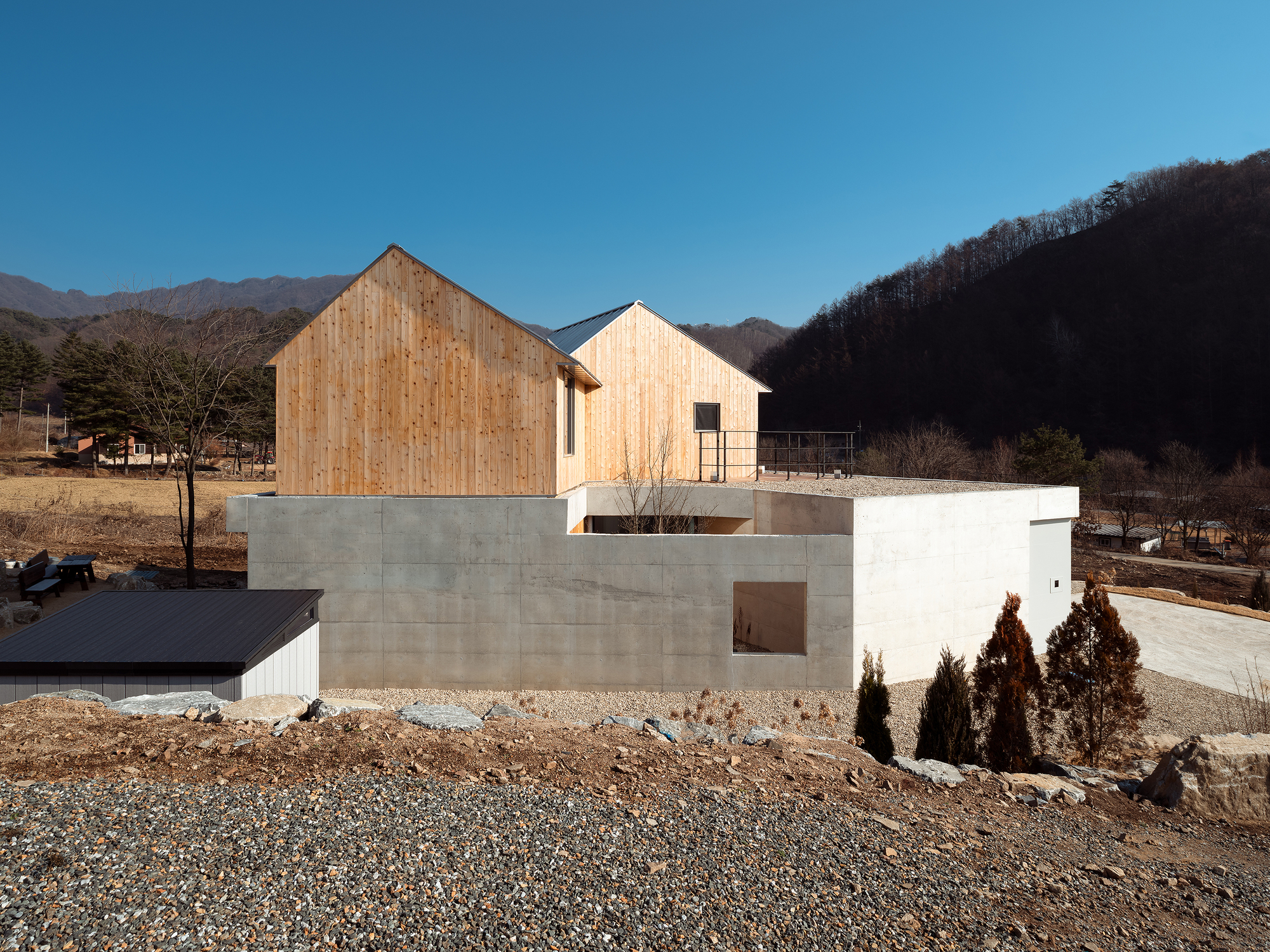 Timber-clad gabled volumes sitting atop a concrete base with gravel site and bare winter hills beyond