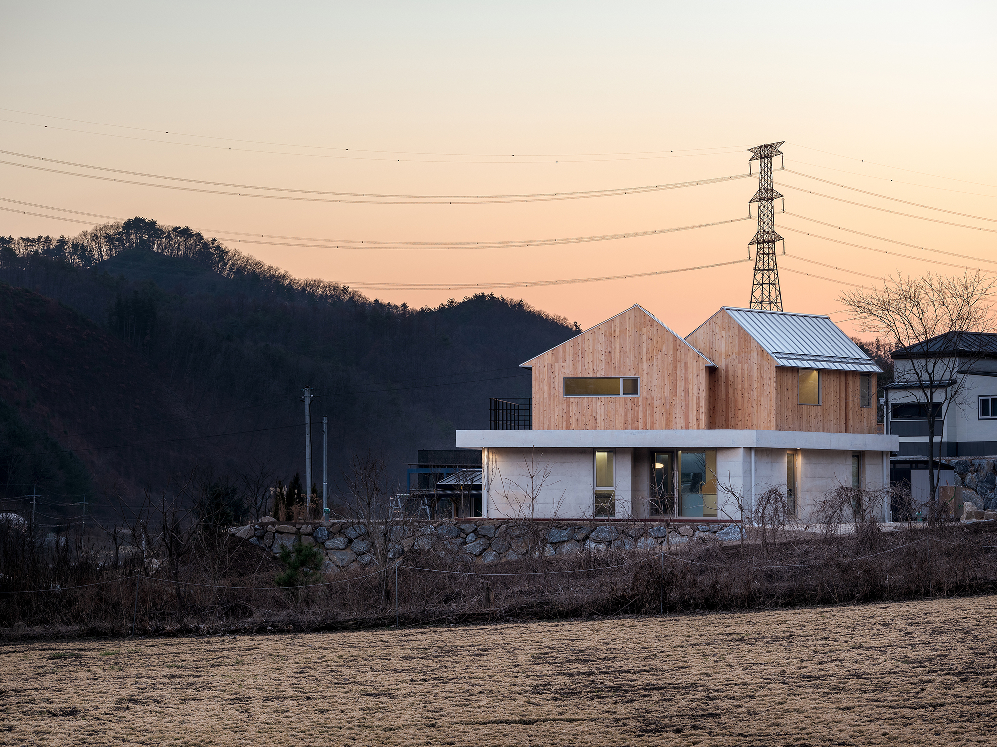 Two-storey residence with timber-clad gabled volumes atop a concrete base at dusk
