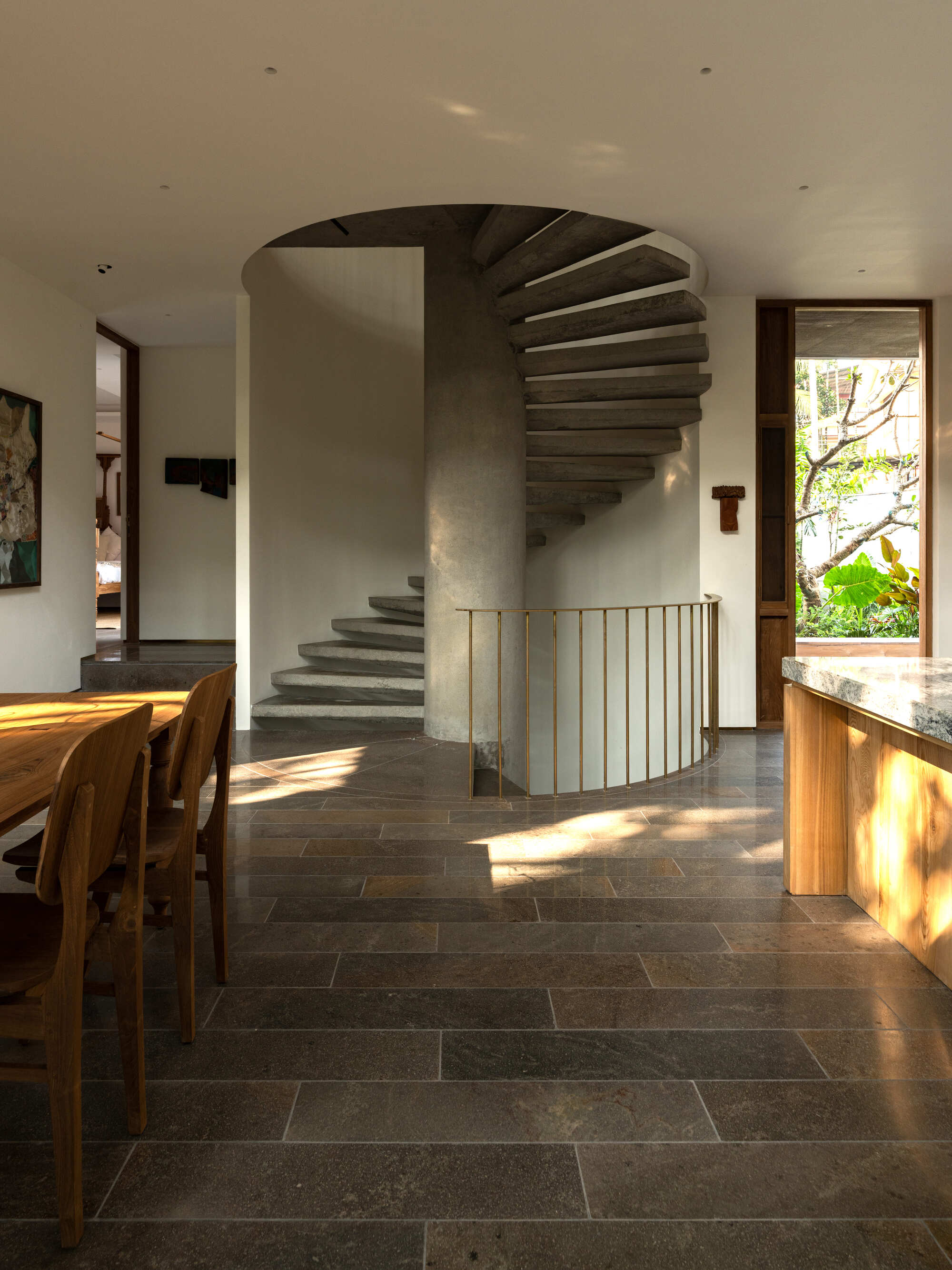 Cylindrical concrete spiral staircase with brass railing in living space with dark tile flooring and afternoon sunlight