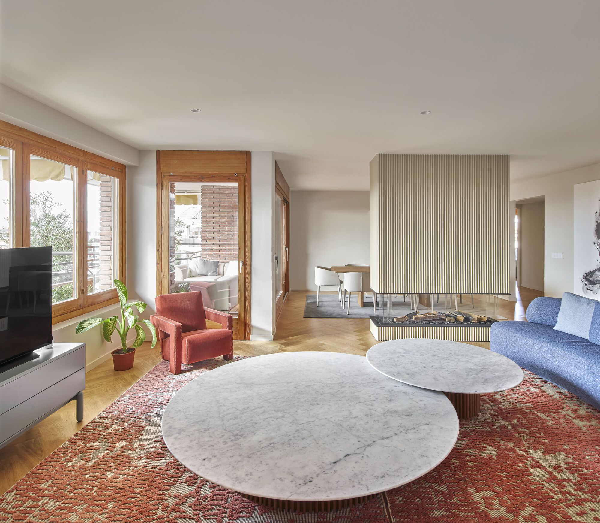 Living room with vertically fluted fireplace wall, marble coffee tables, and timber-framed windows overlooking greenery