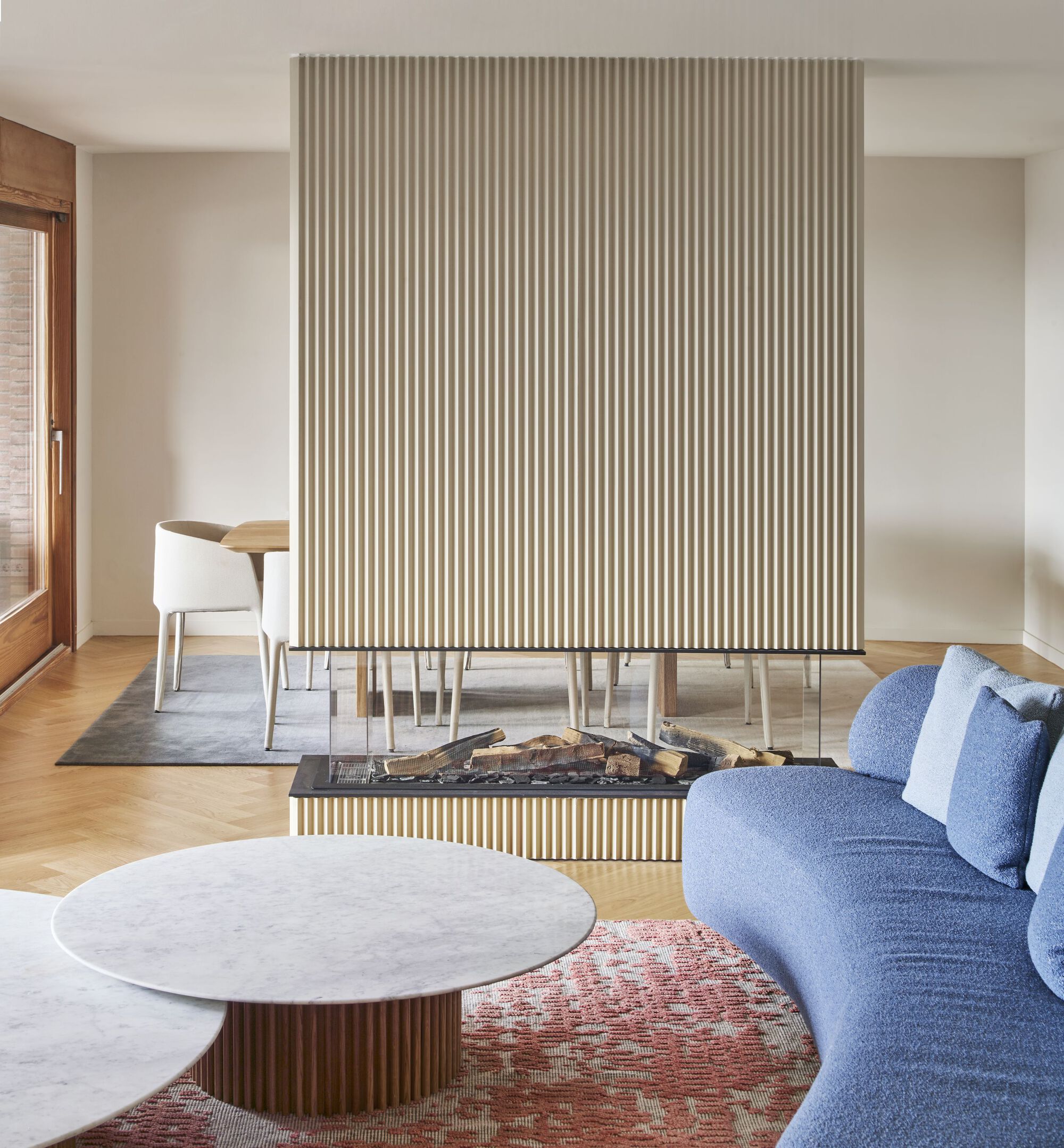 Fireplace with fluted surround holding stacked firewood, flanked by dining table and curved sofa