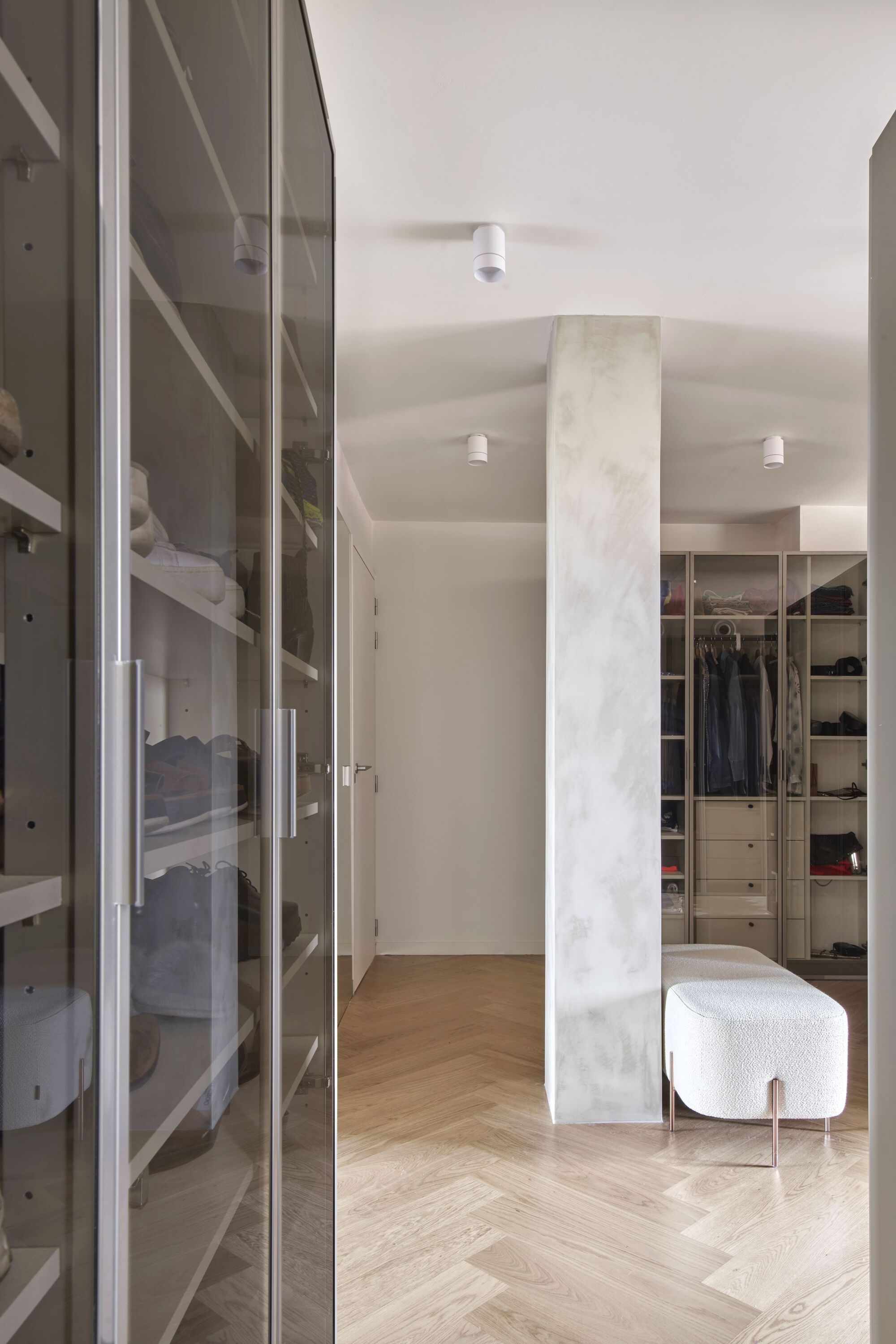 Walk-in closet with glass-fronted storage and white upholstered ottoman on herringbone oak flooring