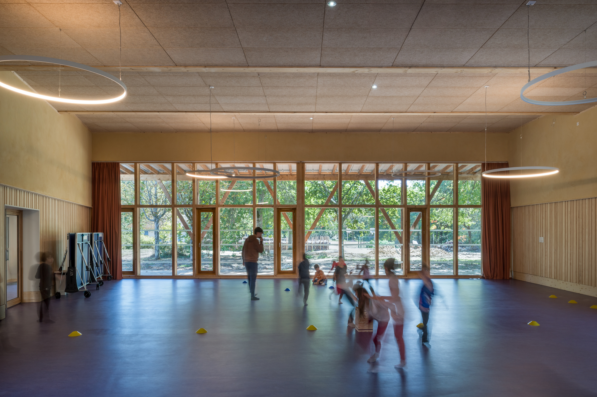 Activity hall with timber-framed glazed wall overlooking trees and children playing on blue flooring