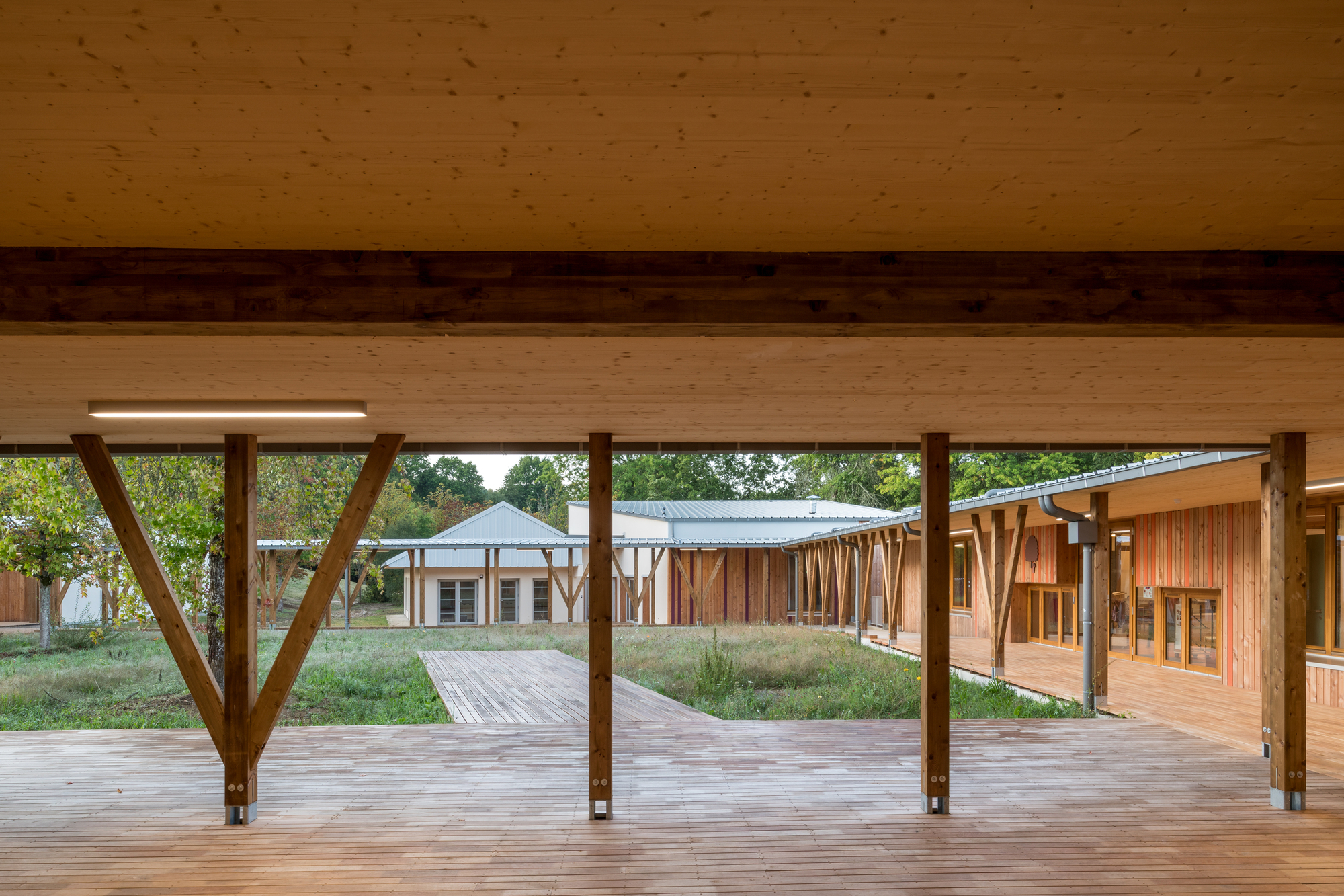 Covered timber walkway with diagonal columns framing views across a planted courtyard to adjacent timber structures