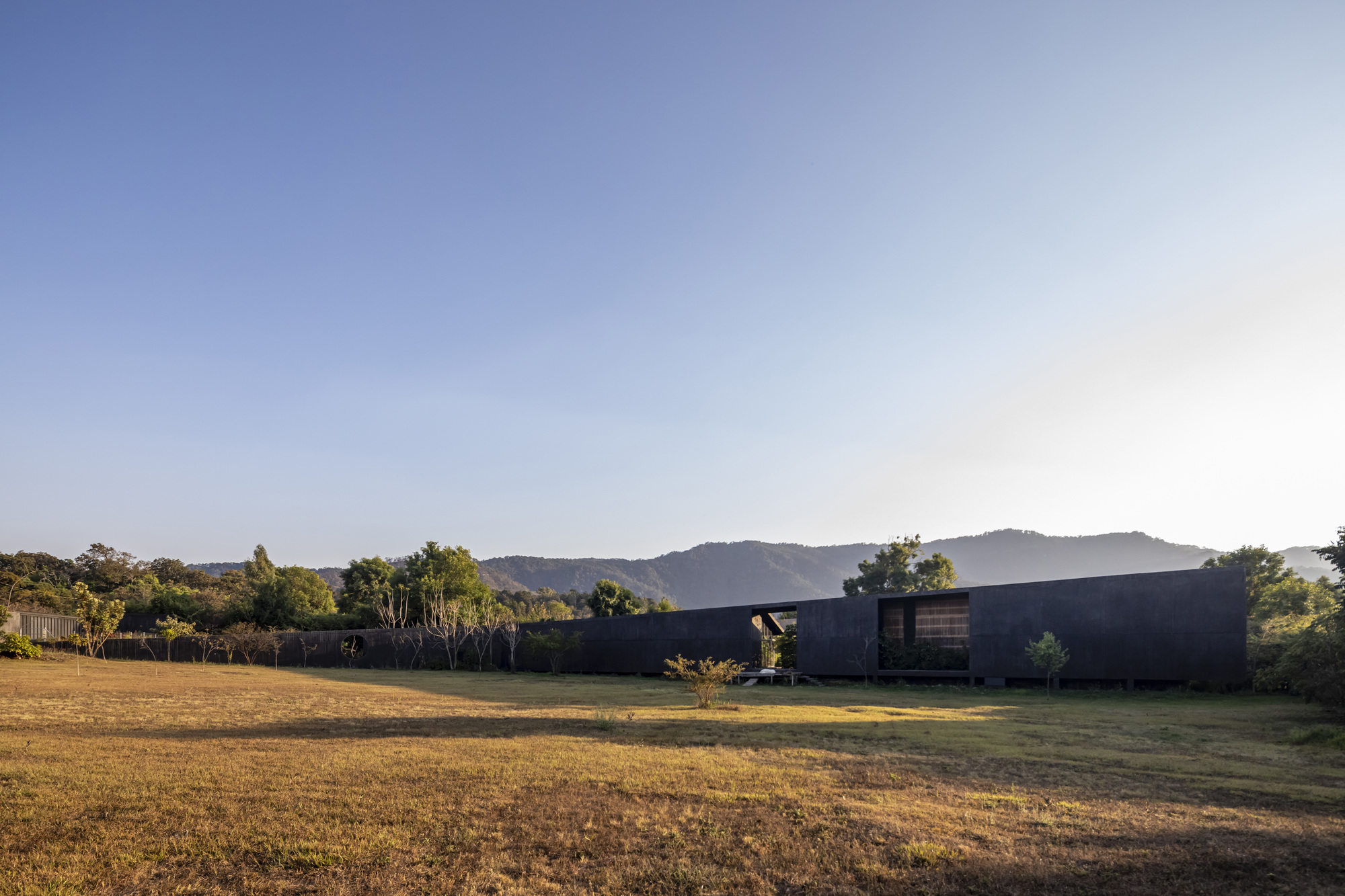 Long concrete facade with small window openings facing a dry grass field at dusk