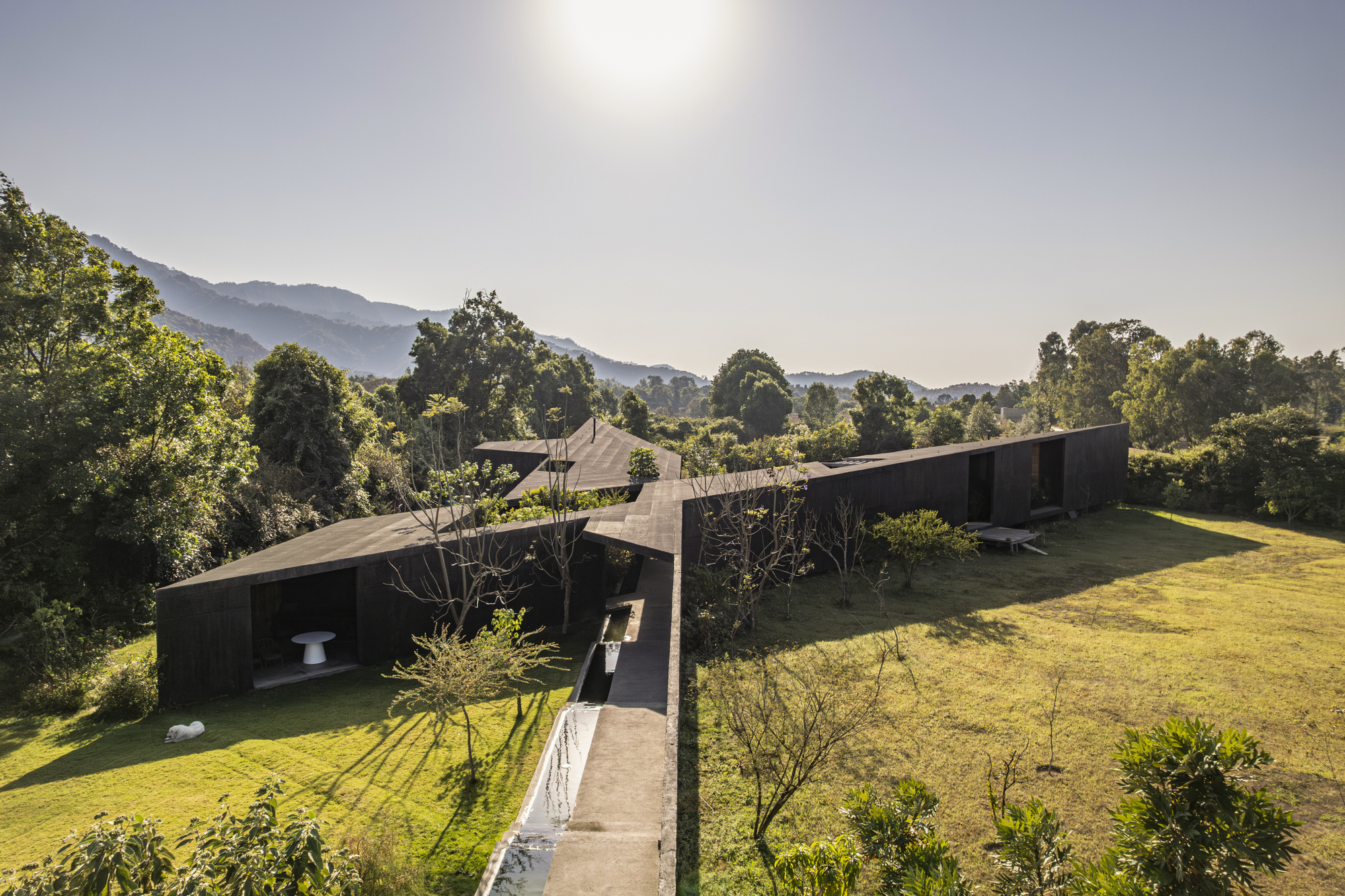 Aerial view of linear dark-clad volumes connected by a concrete bridge spanning a sloped lawn with mountains beyond