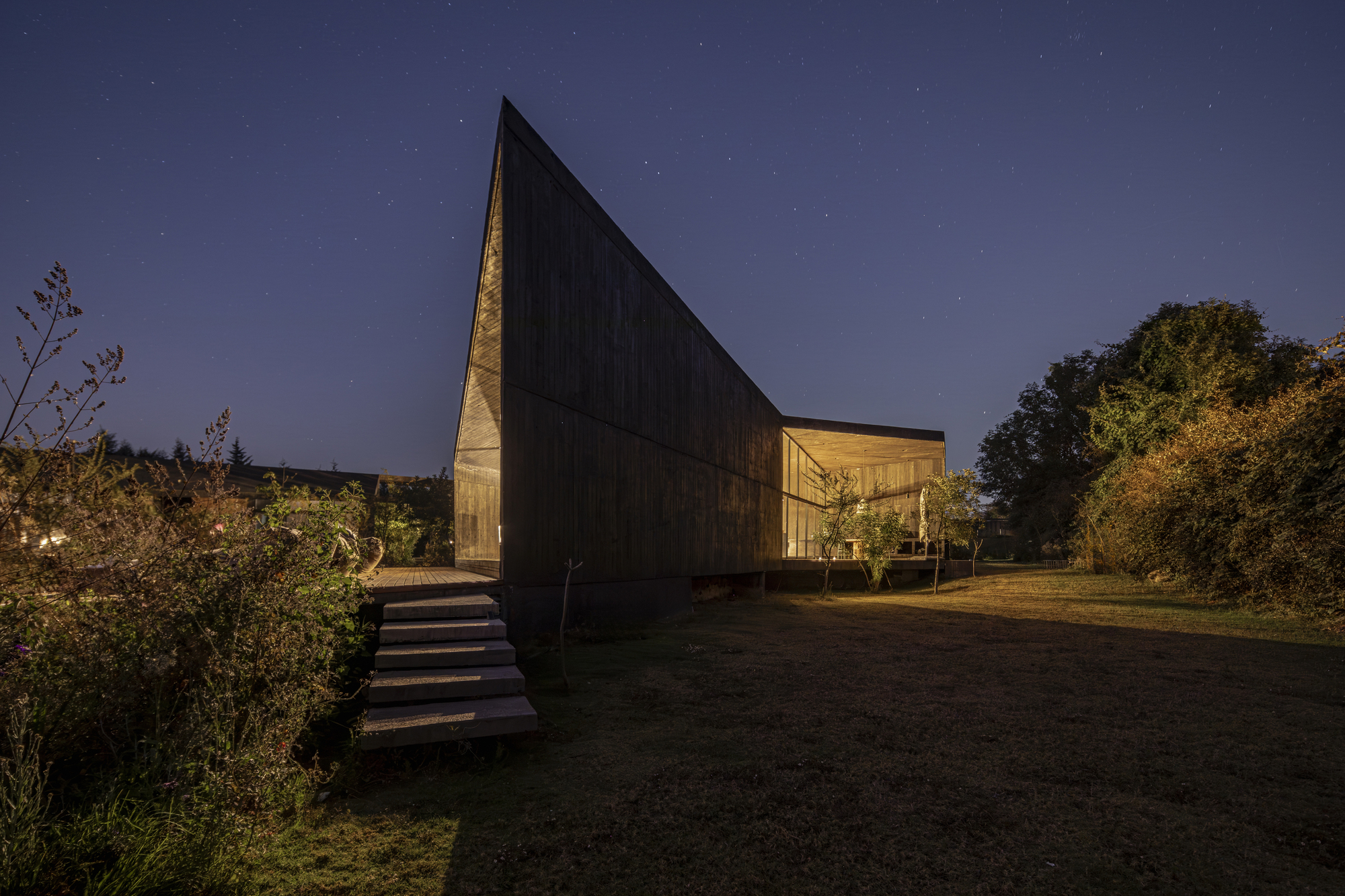 Elevated dark timber volume with illuminated glass pavilion beside it under a starlit night sky