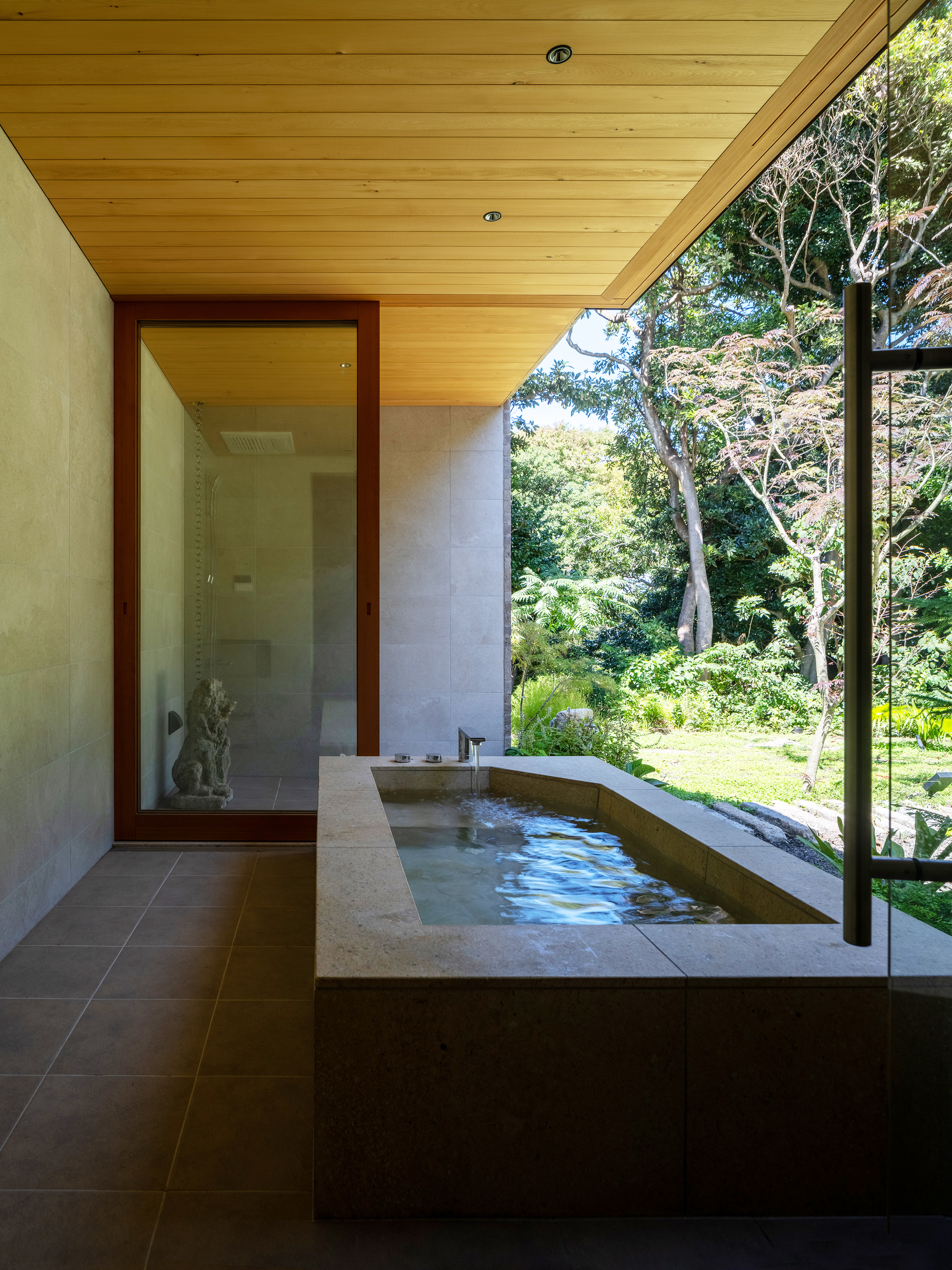 Bathroom with sunken concrete tub beside floor-to-ceiling glass overlooking garden and trees