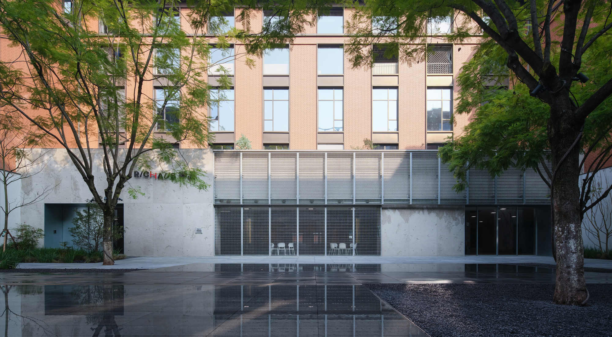 Courtyard facade with horizontal metal louvers reflecting in a dark water pool flanked by trees