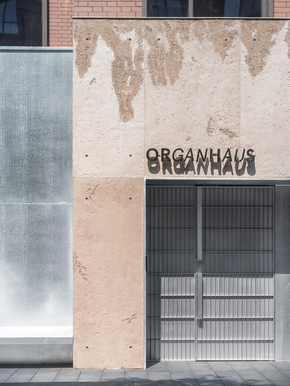 Weathered rammed earth facade with textured layering and metal screen window opening below signage