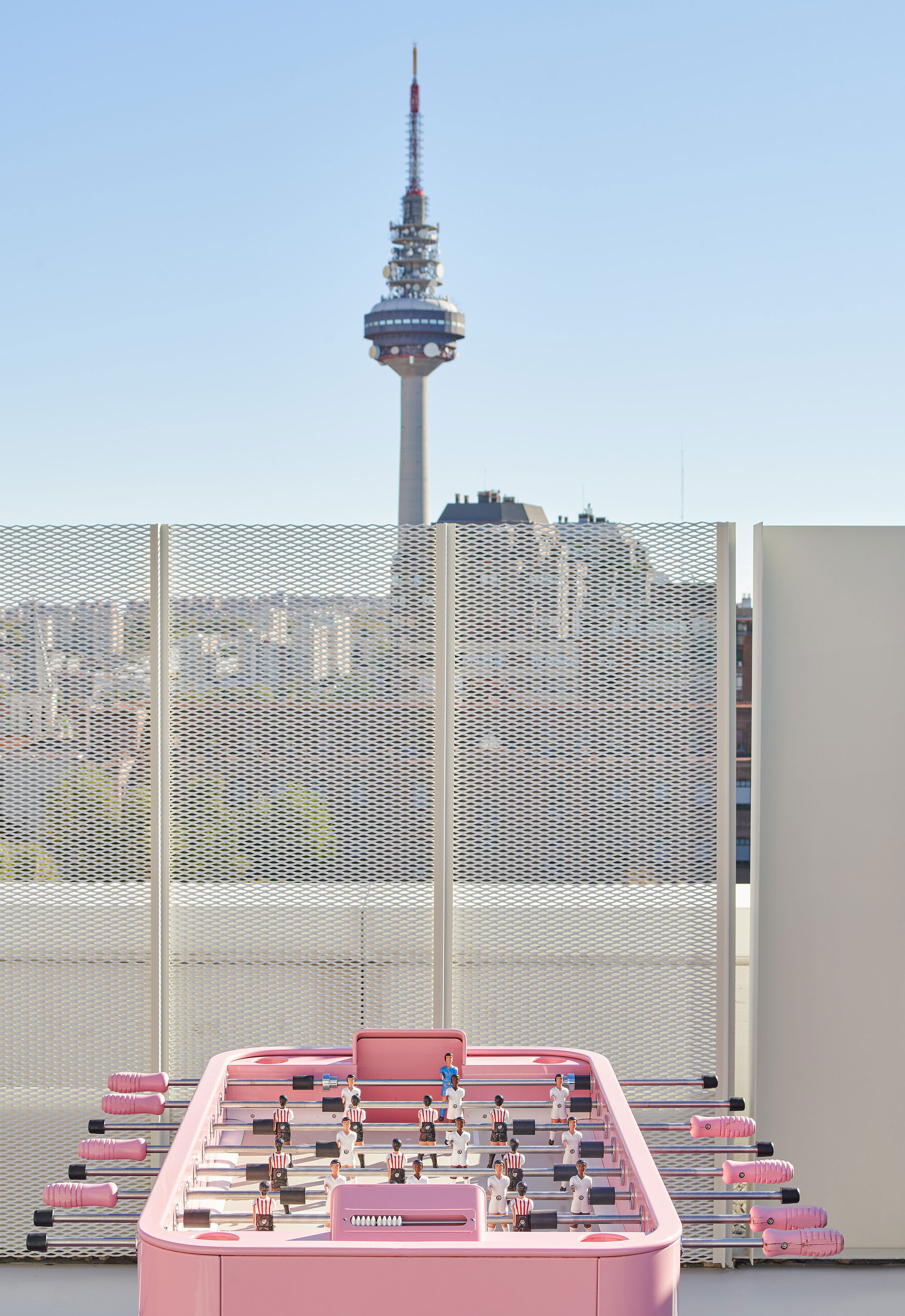 Pink foosball table on rooftop with perforated metal screens and telecommunications tower in distance