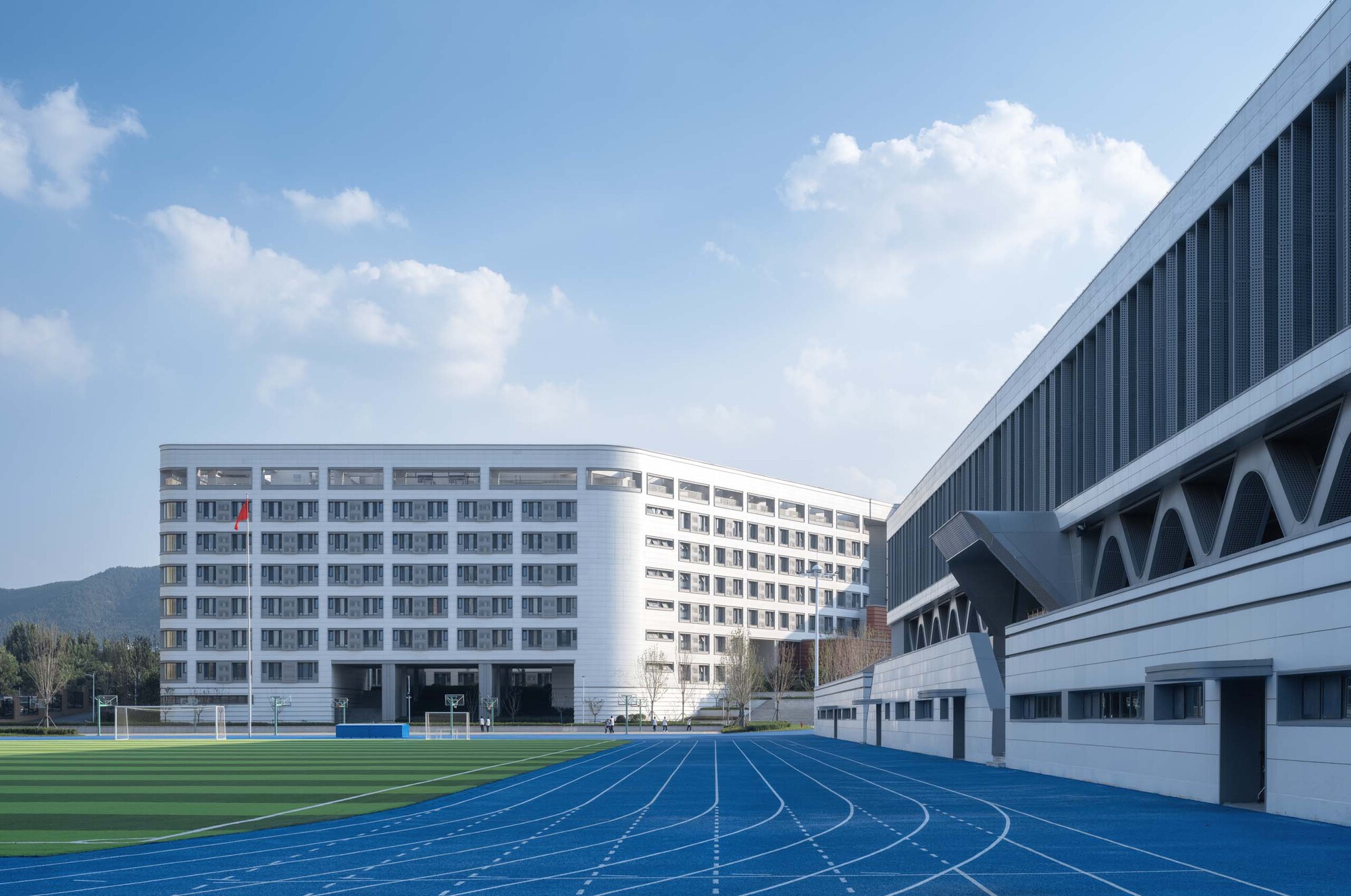 Campus view showing white residential towers and gymnasium with diagonal bracing beyond a blue running track