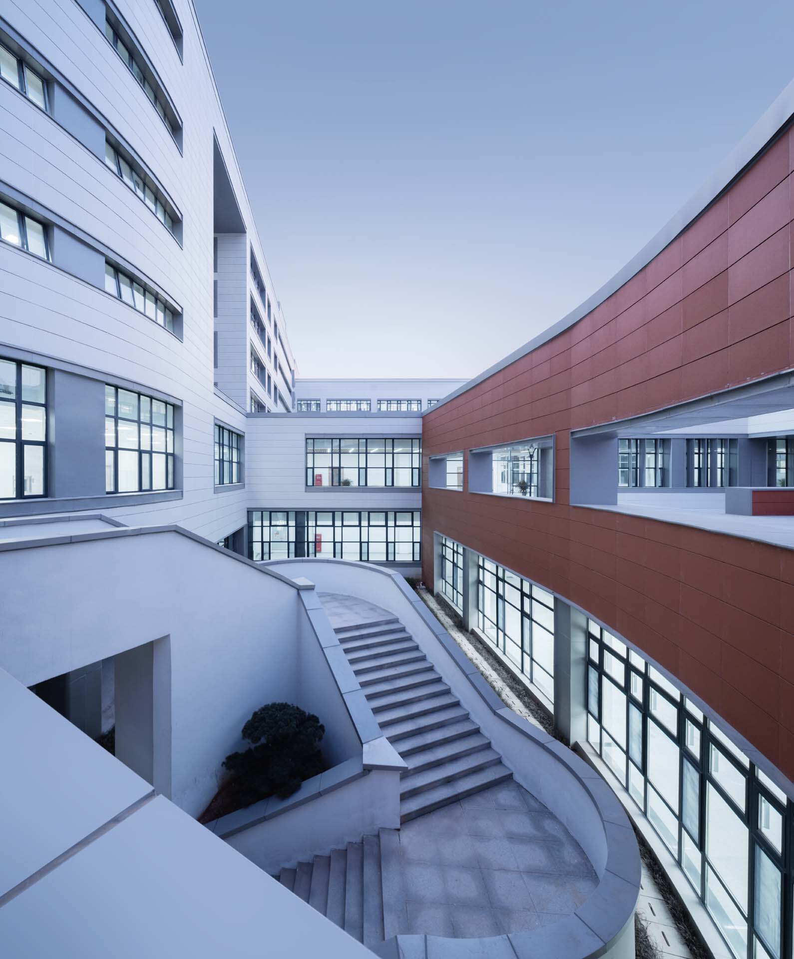 Curved staircase descending into a sunken courtyard between white and terracotta panel facades with glazed bridges
