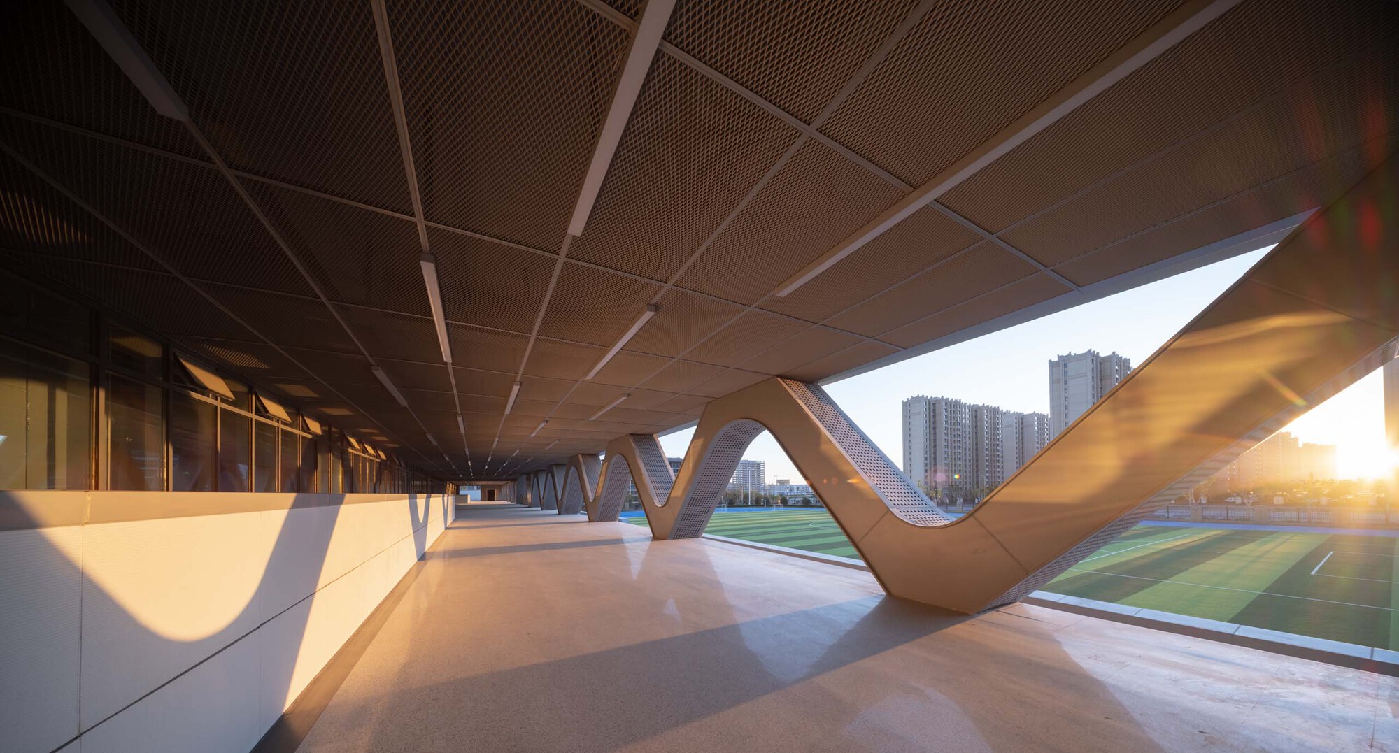 Covered colonnade with V-shaped columns and perforated wood ceiling panels at sunset