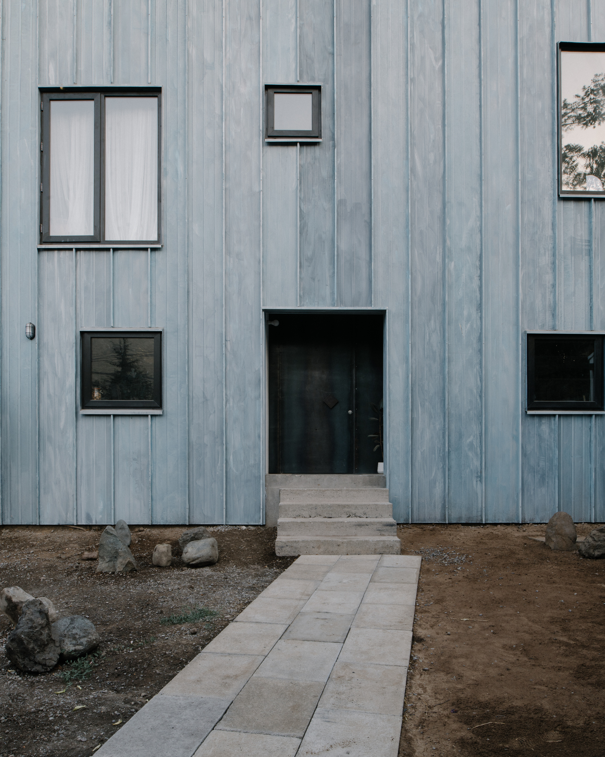 Weathered vertical timber facade with black-framed windows and door, concrete pavers leading to entrance