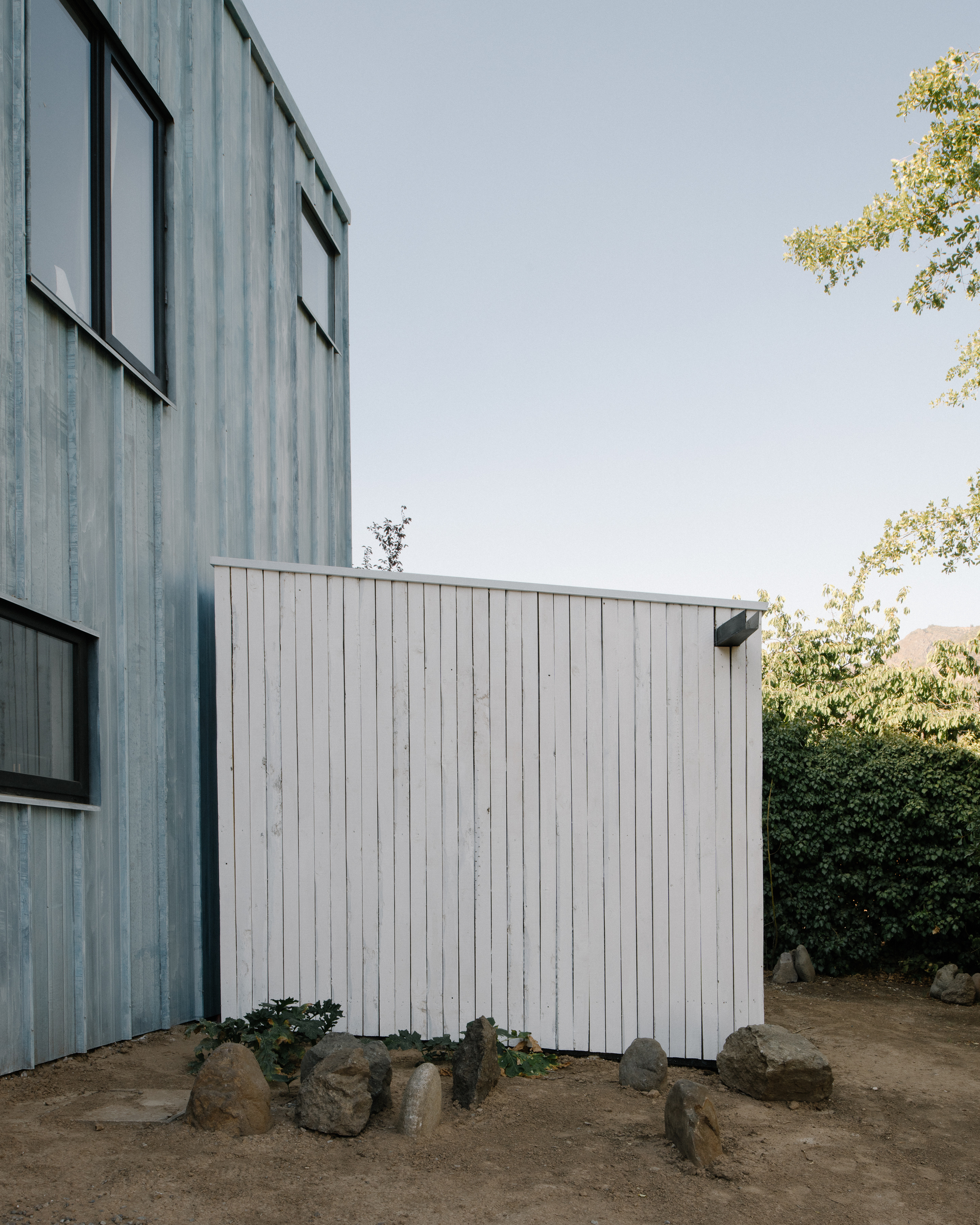 Junction of weathered blue-grey and white vertical timber volumes with rocks and vegetation at base