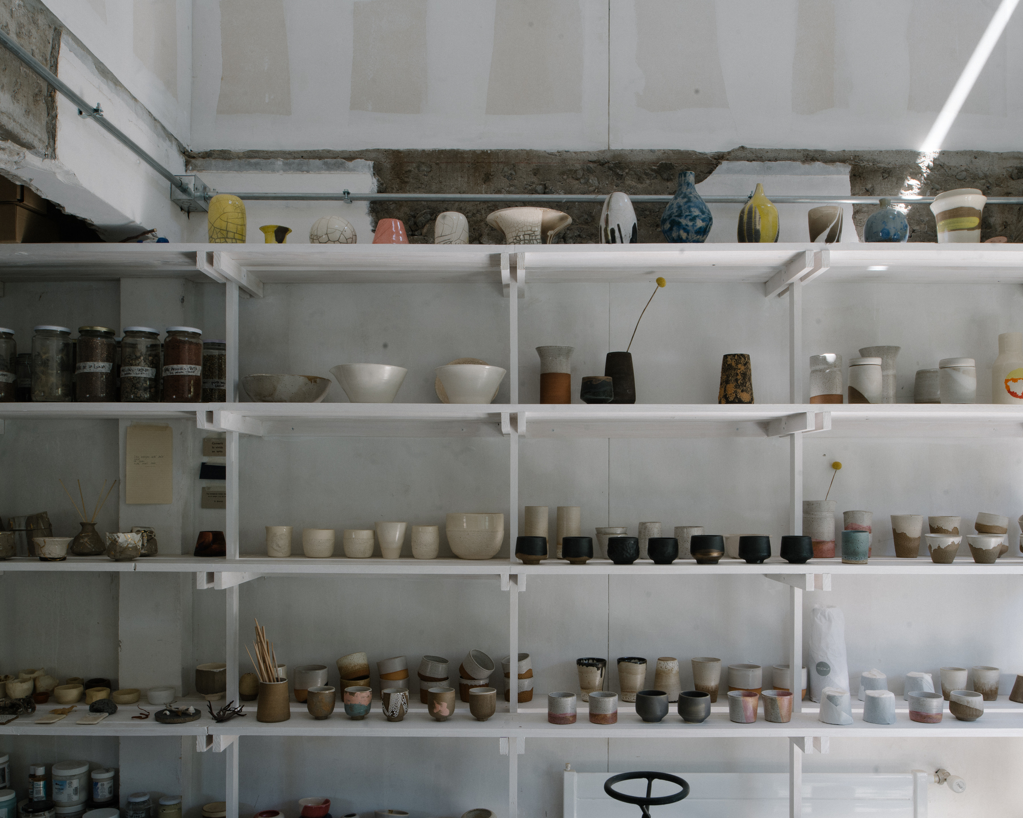 White metal shelving filled with ceramic vessels and pottery beneath a raw concrete beam