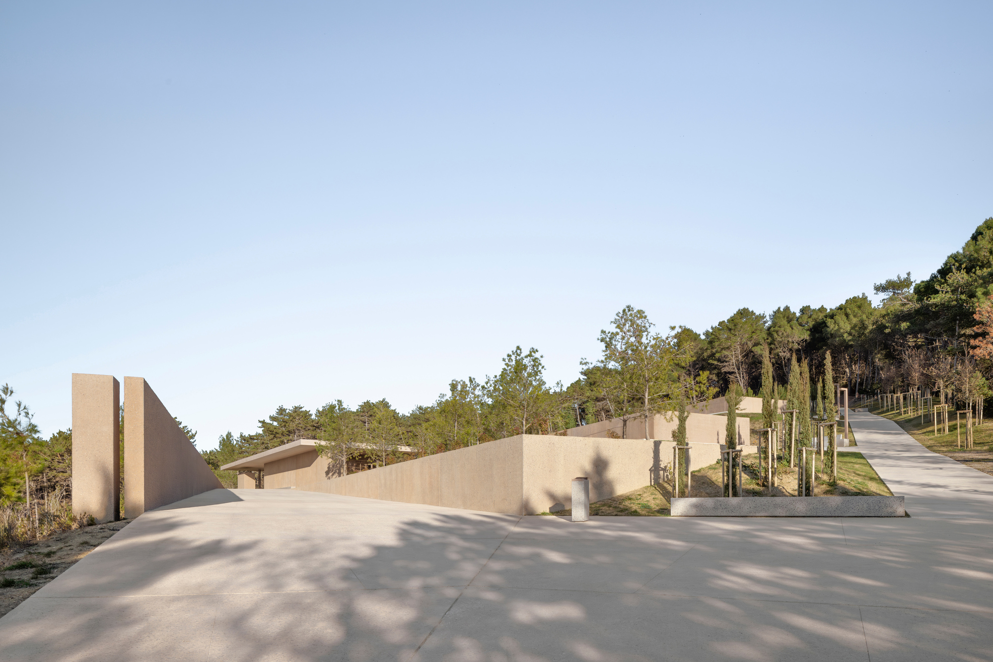 Concrete plaza with angular walls and grouped timber posts among shadows cast by surrounding trees