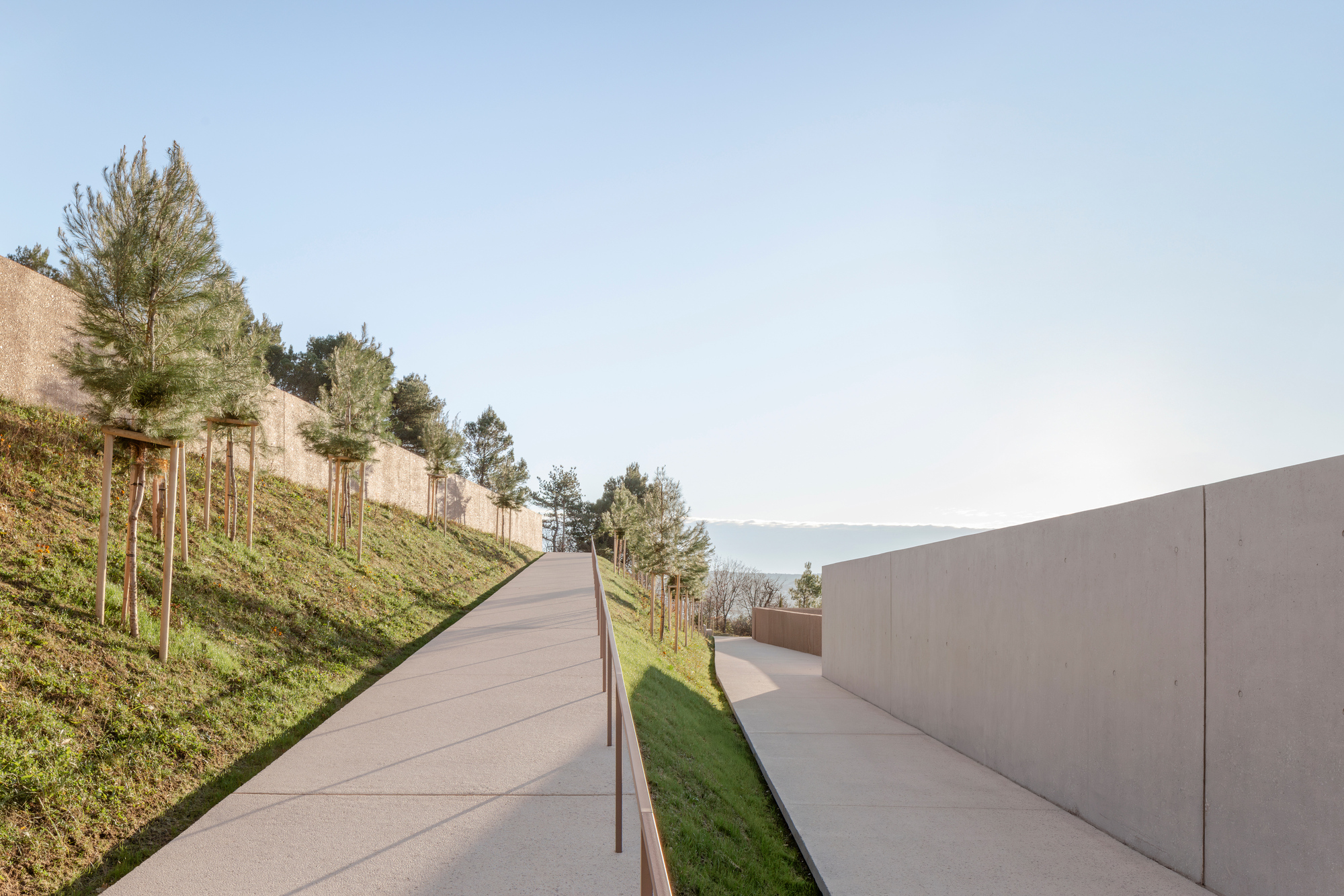 Concrete ramp ascending hillside between planted slope and smooth retaining wall with distant lake