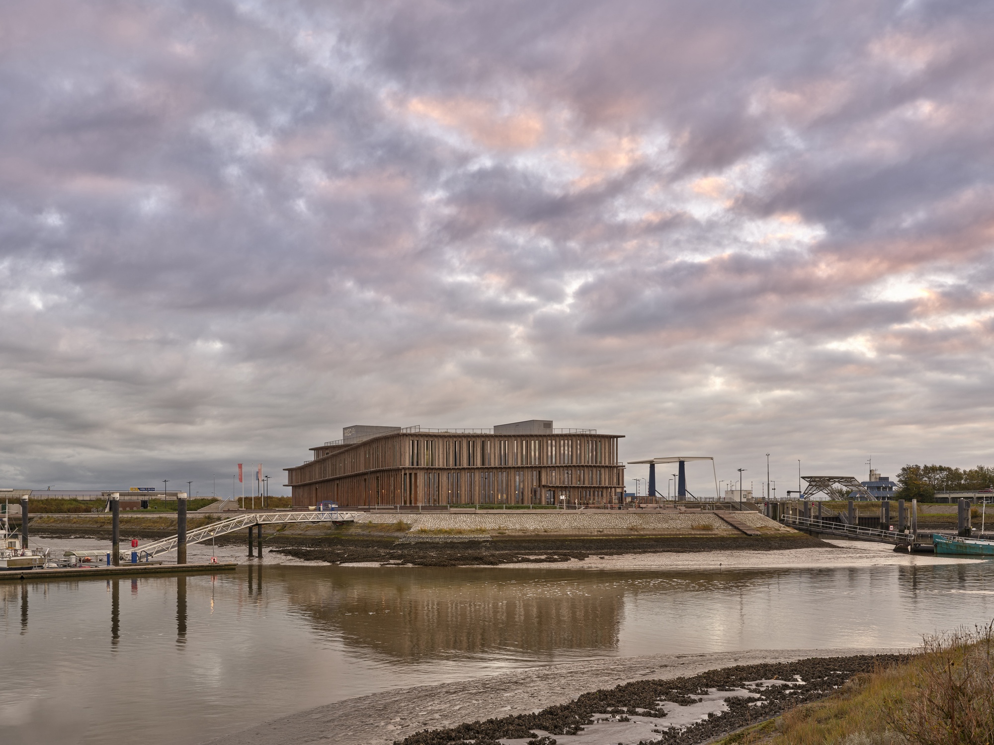 Horizontal timber-clad building reflected in the tidal waters of a mudflat harbor under dramatic clouds