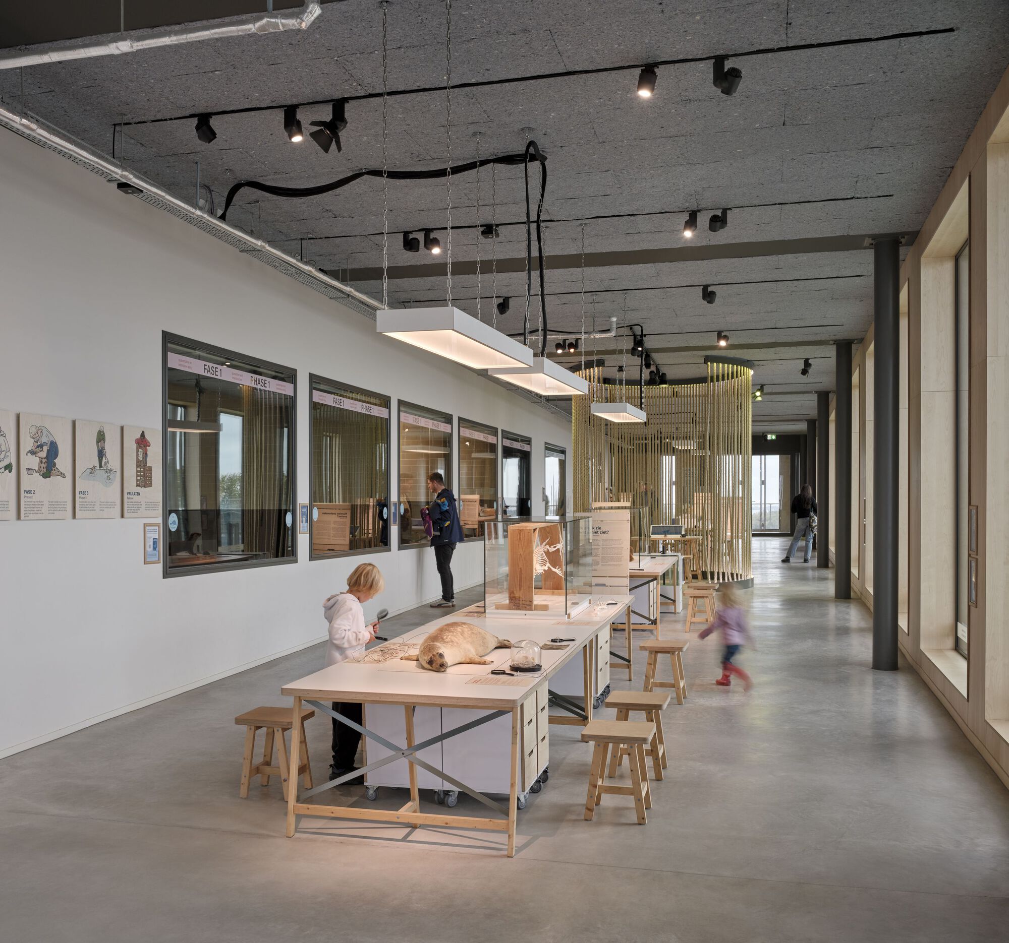 Exhibition hall with concrete plank ceiling, work tables, wooden stools, and children engaging with displays