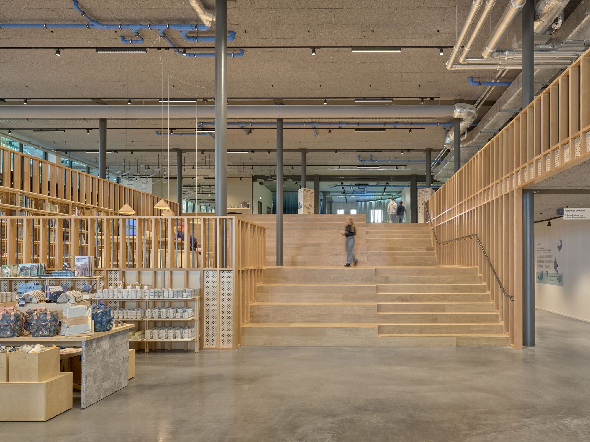 Wide timber staircase ascending past a retail display wall under an exposed concrete soffit with mechanical ducts