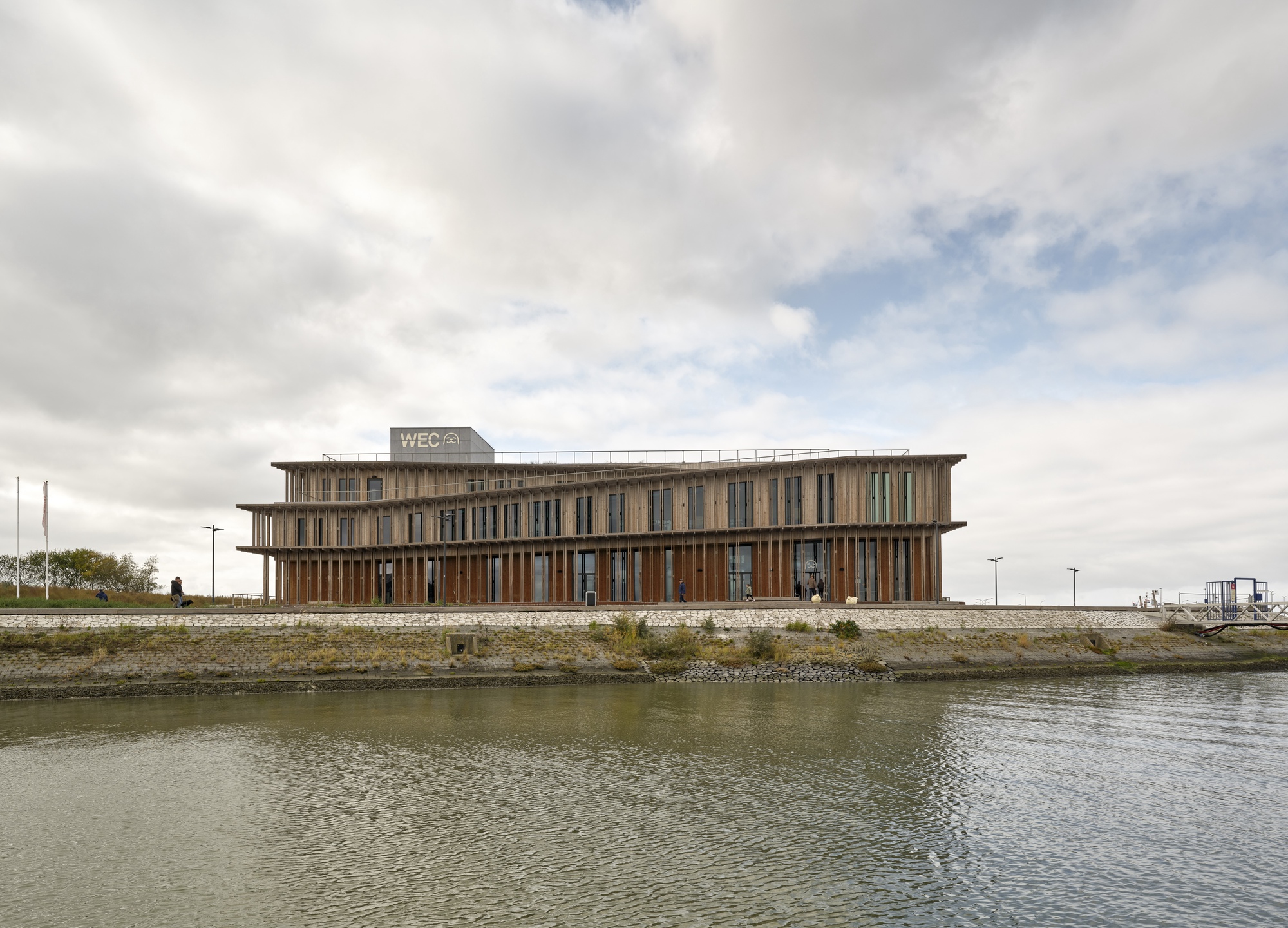 View across the canal showing the elevated building with timber brise-soleil on a stone plinth