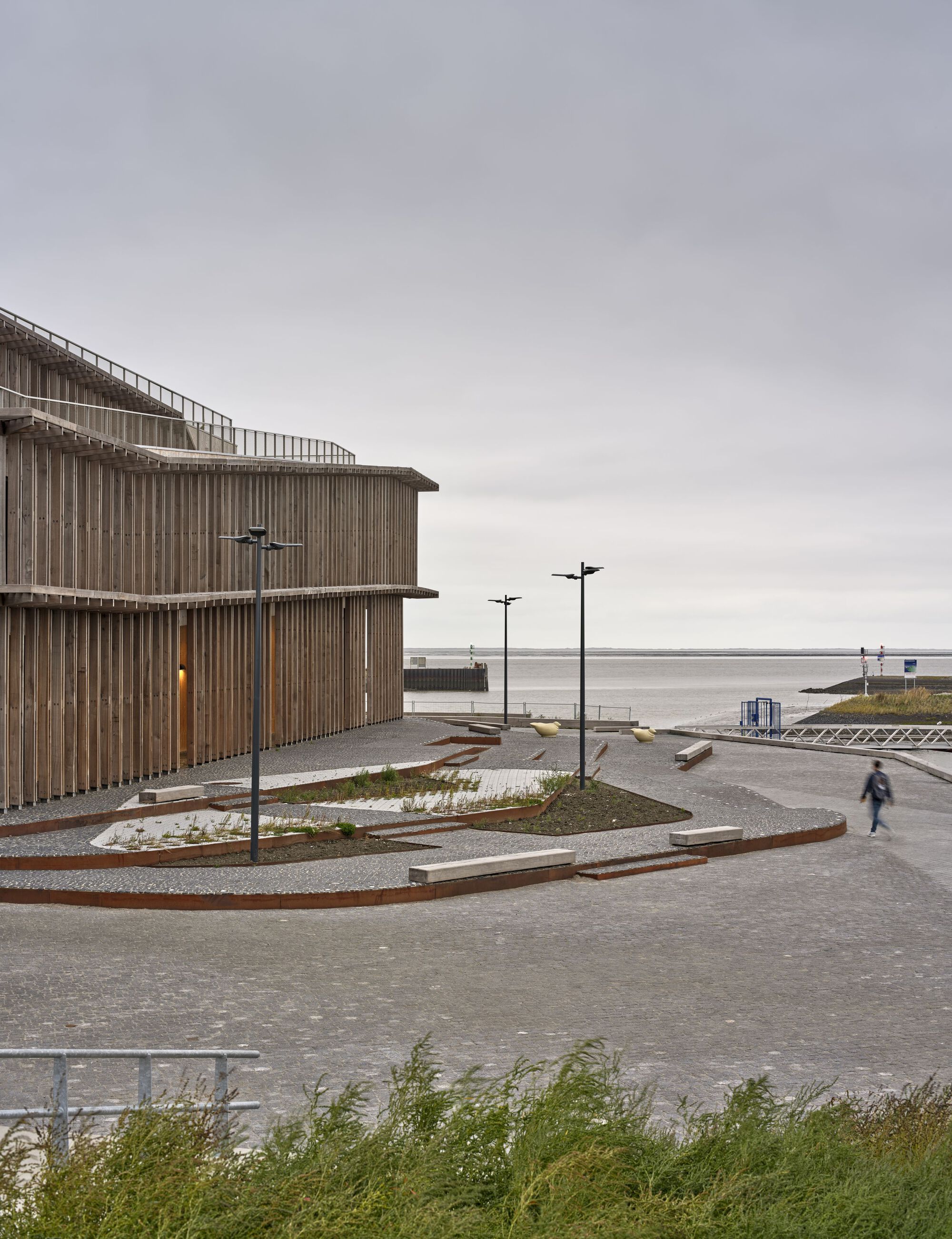 View of the curved timber facade with terraced paving and a lone figure walking along the waterfront