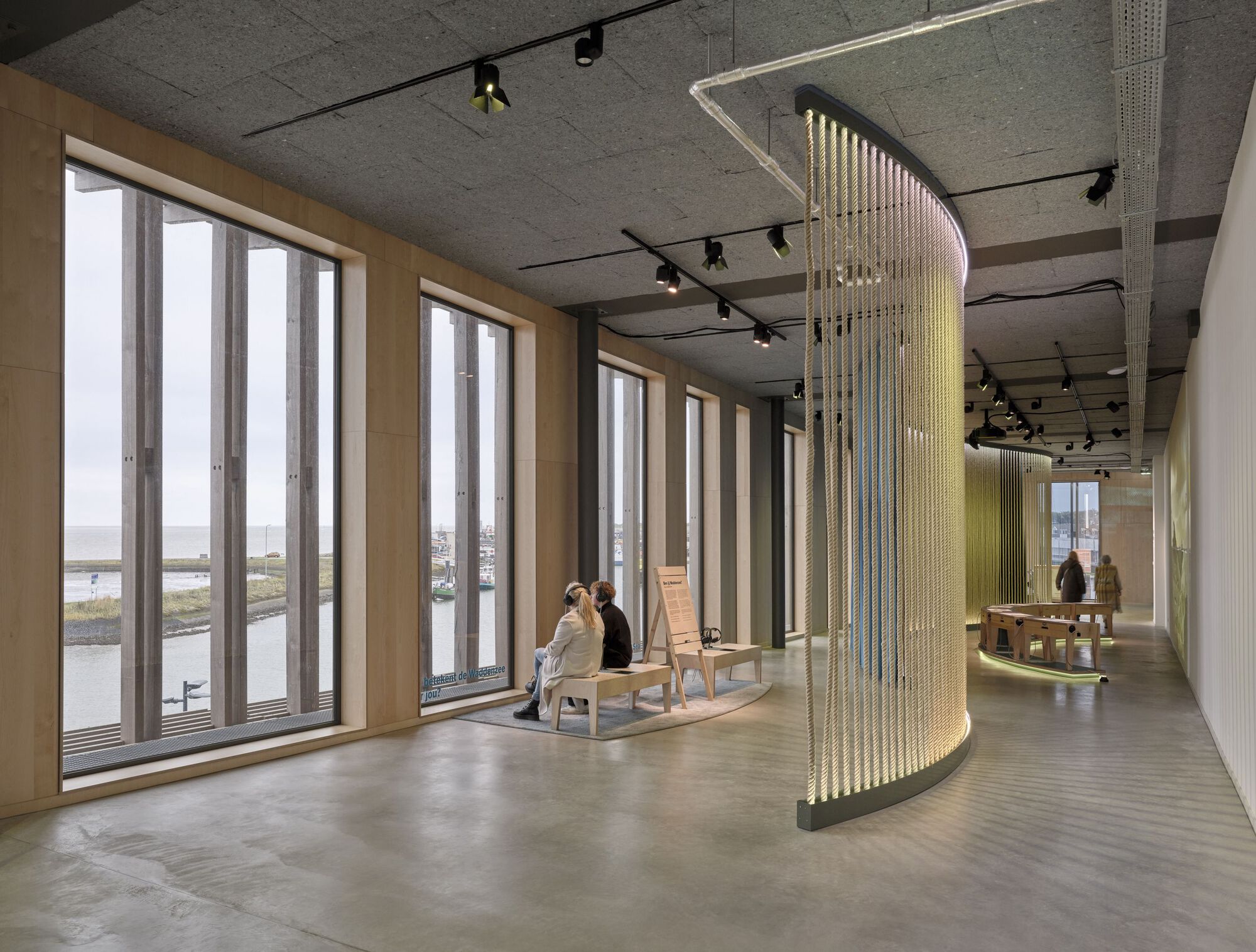 Curved brass bead curtain installation beside tall timber-framed windows with person seated near waterfront view