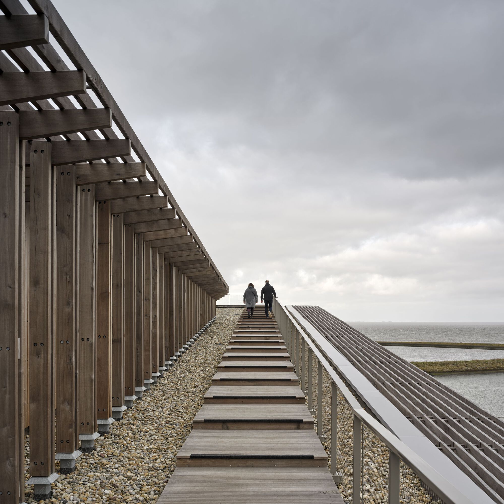 Timber boardwalk with open stairway alongside vertical slat screen and gravel ballast roof overlooking the sea