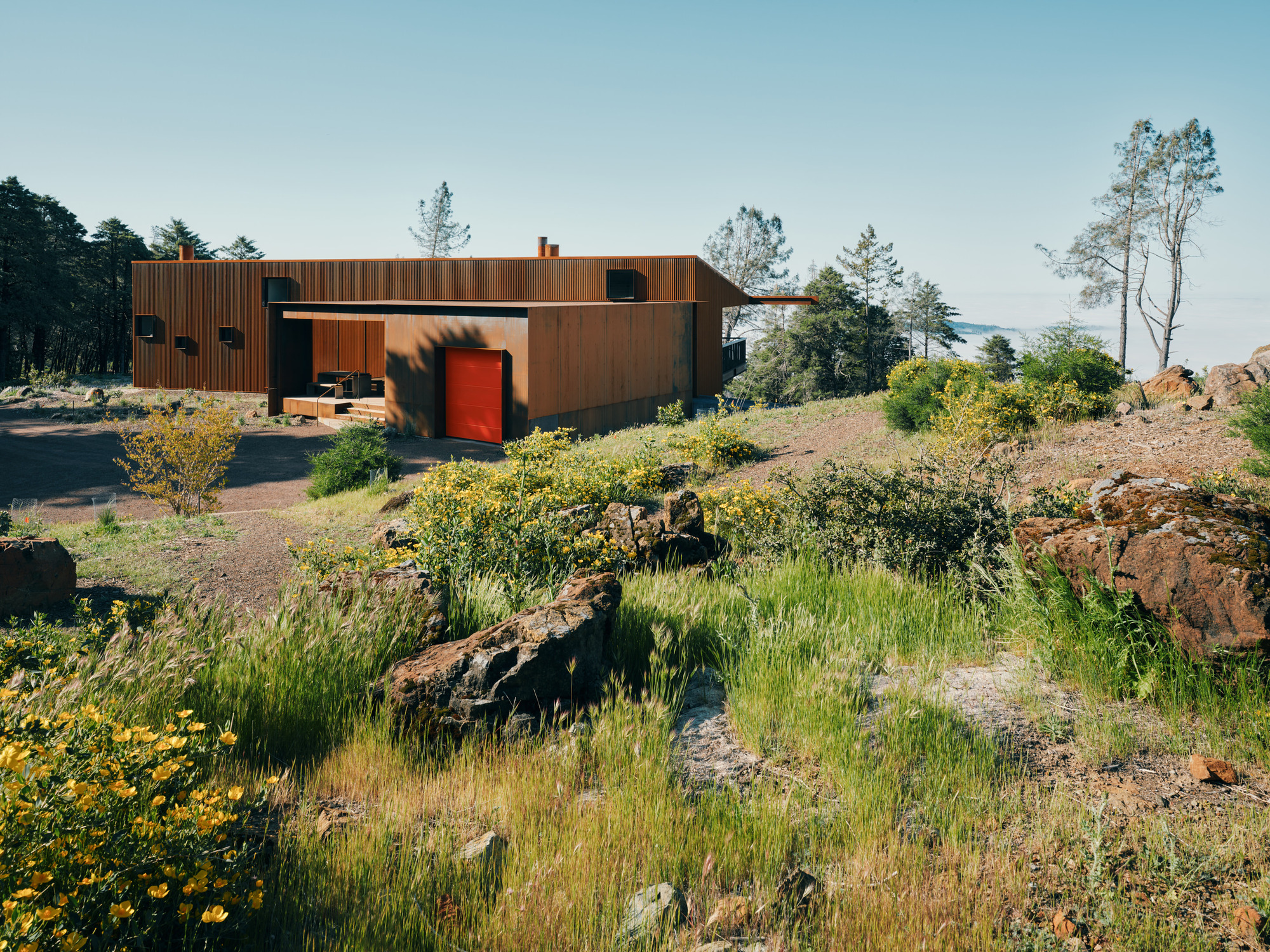 Weathered steel and timber residence with red entry door set among wildflowers and volcanic rock outcroppings