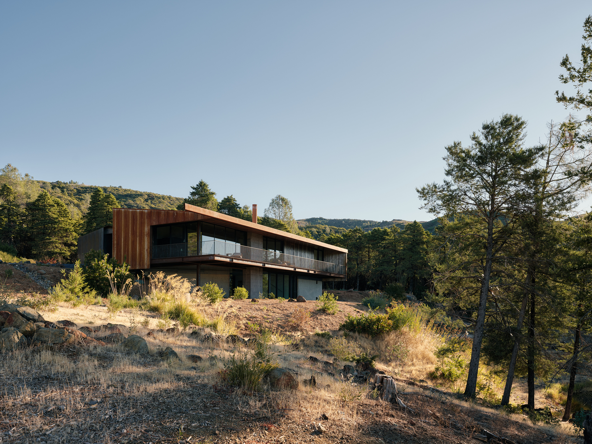 Sloped metal roof over cantilevered volume on a rocky hillside with sparse vegetation and distant ridges