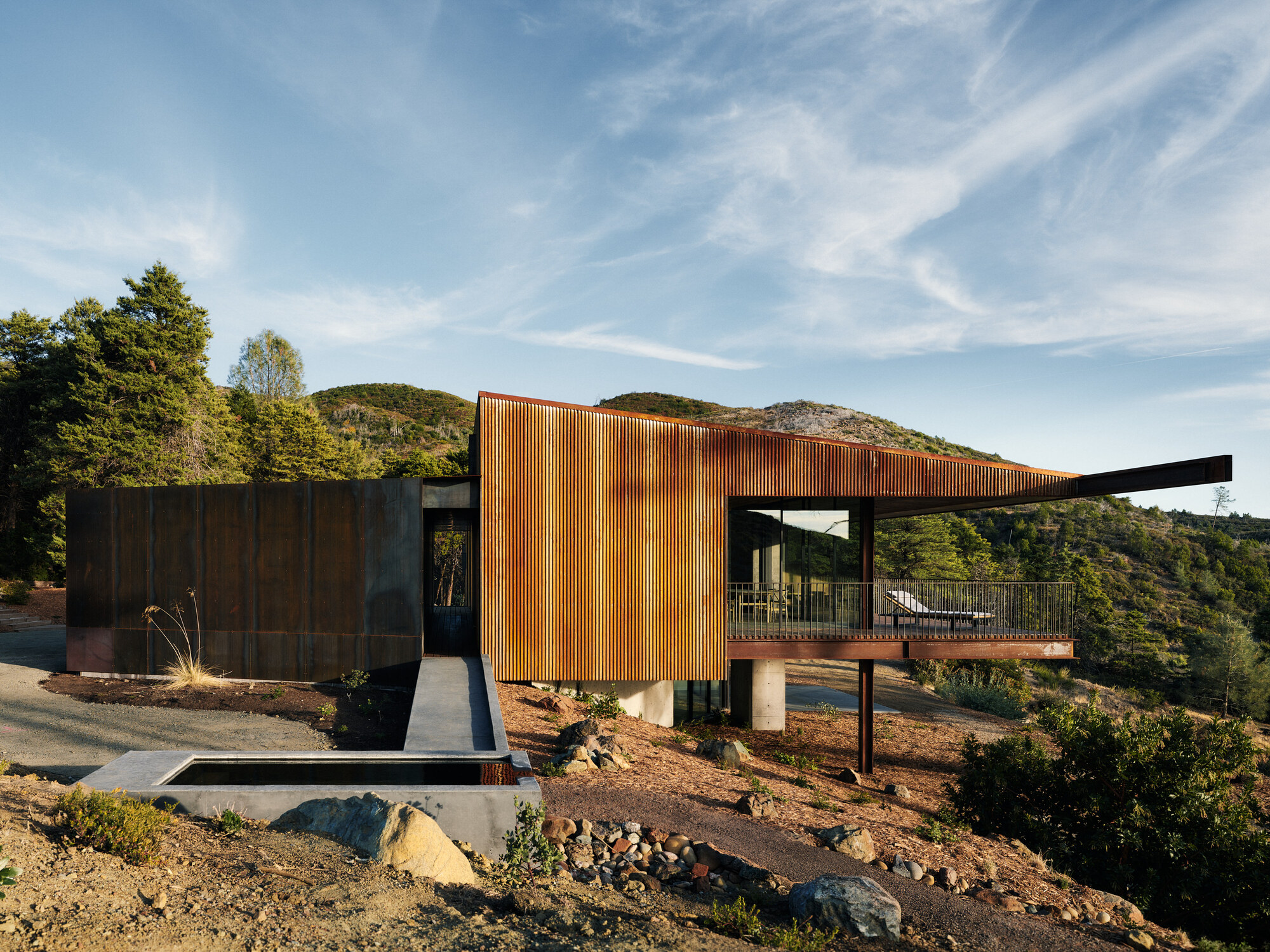 Corrugated metal facade with vertical fins next to weathered steel panels on pilotis above exposed bedrock