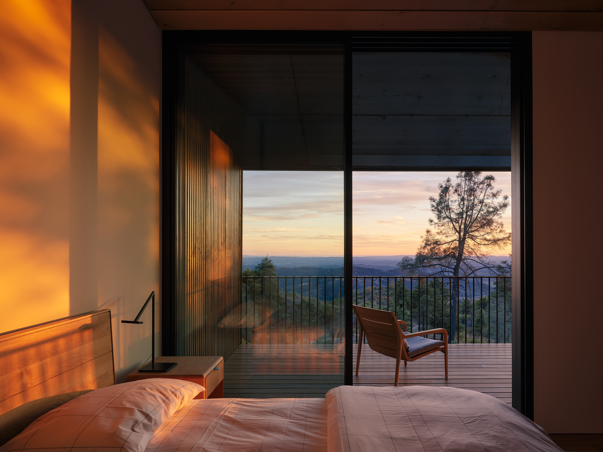 Bedroom with glazed doors opening to a timber deck overlooking distant hills at sunset