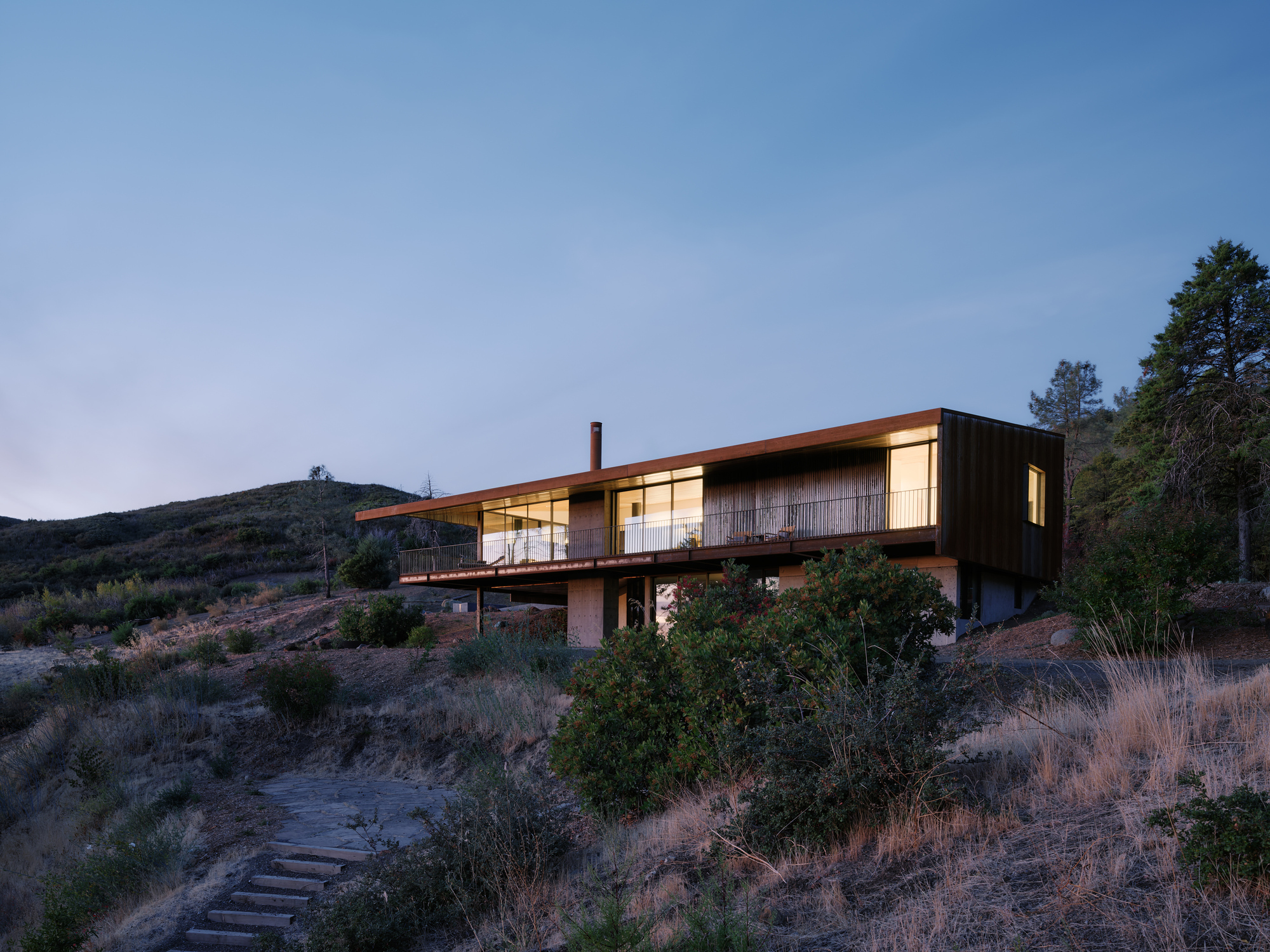 Two-level residence with corten upper volume and timber lower level nestled in scrubland at twilight