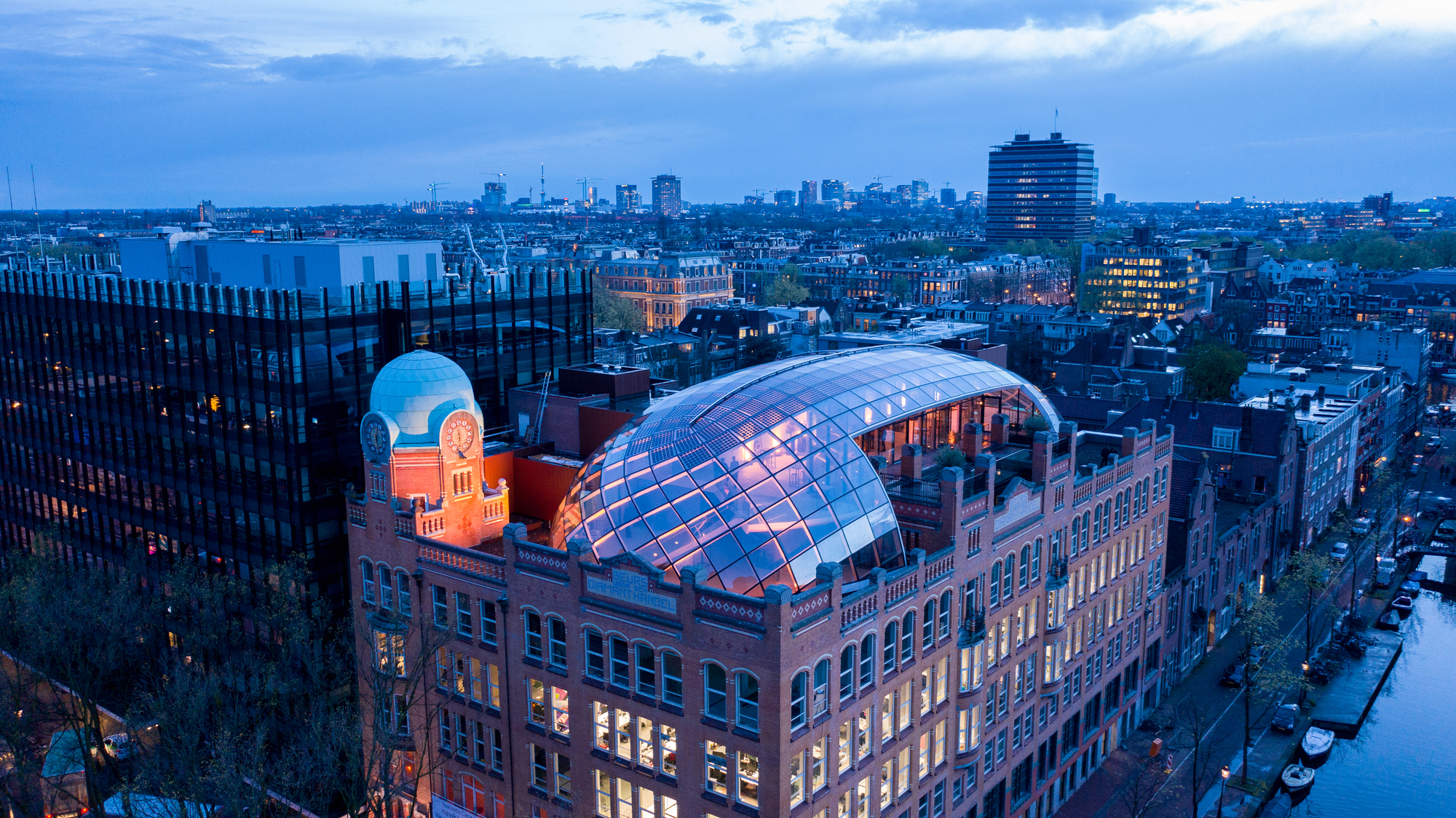 Illuminated glass dome rising from a brick building at twilight with cityscape beyond
