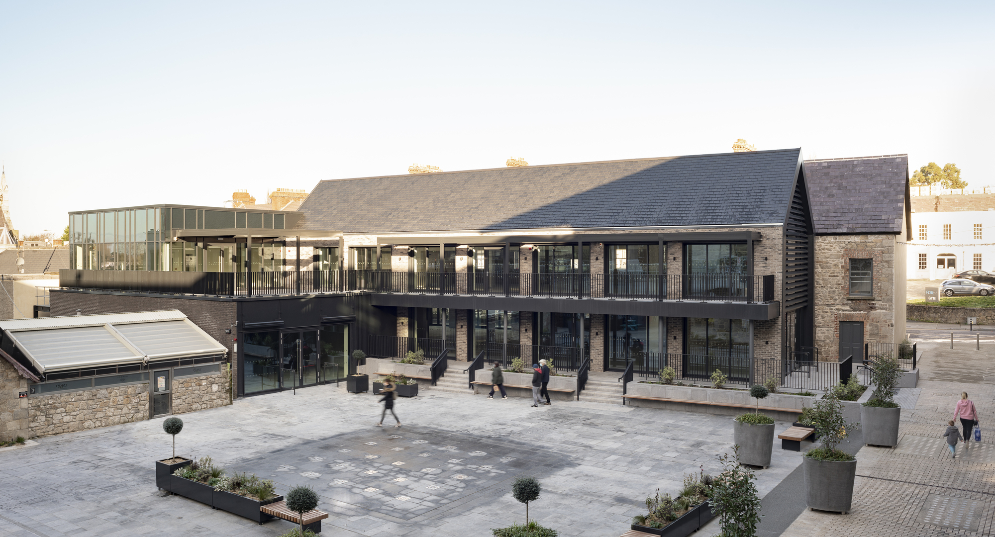 Public courtyard with grey paving and planted containers between restored brick buildings and glazed volumes