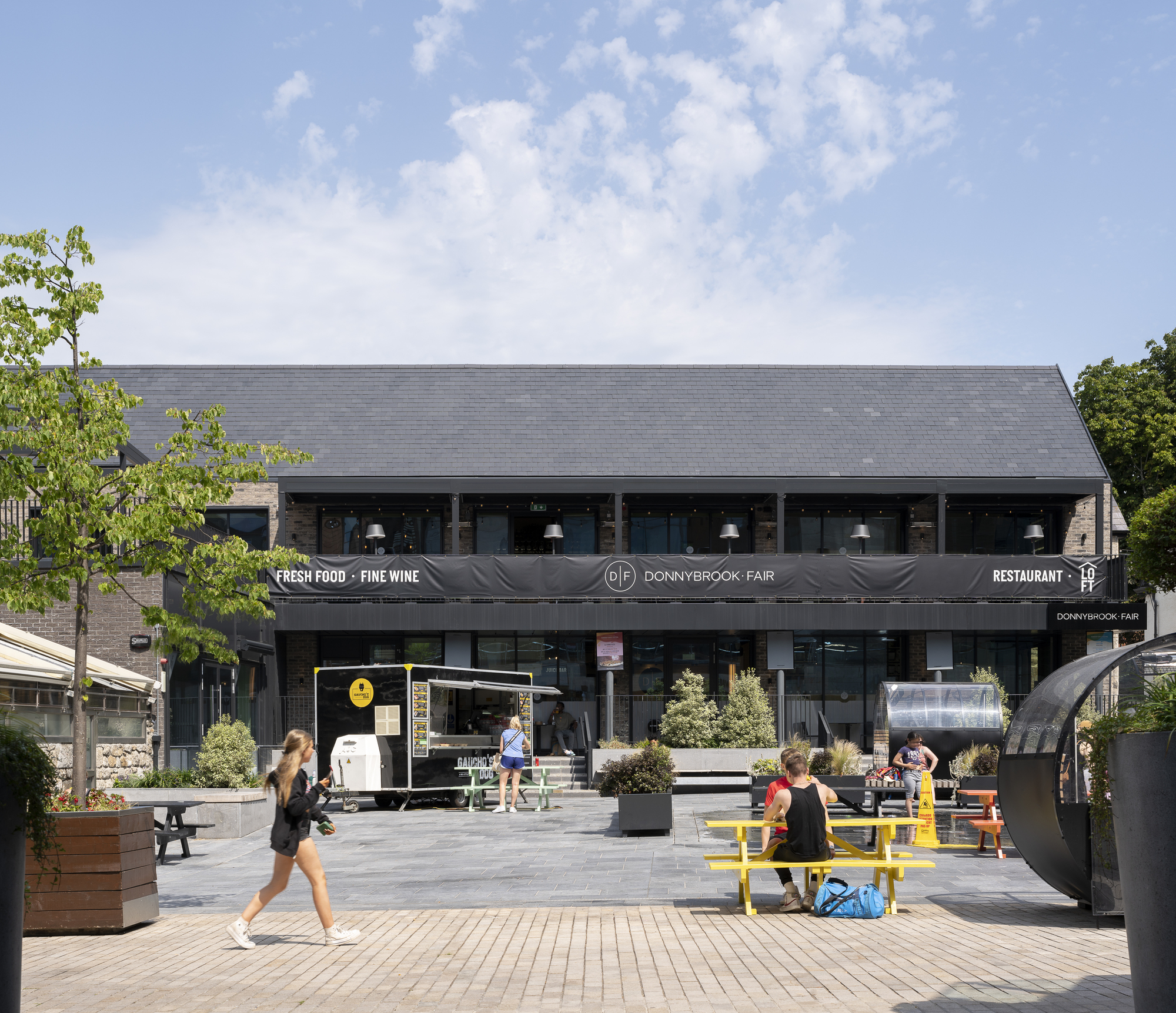 Public plaza with yellow picnic tables and pedestrians in front of two-story commercial building