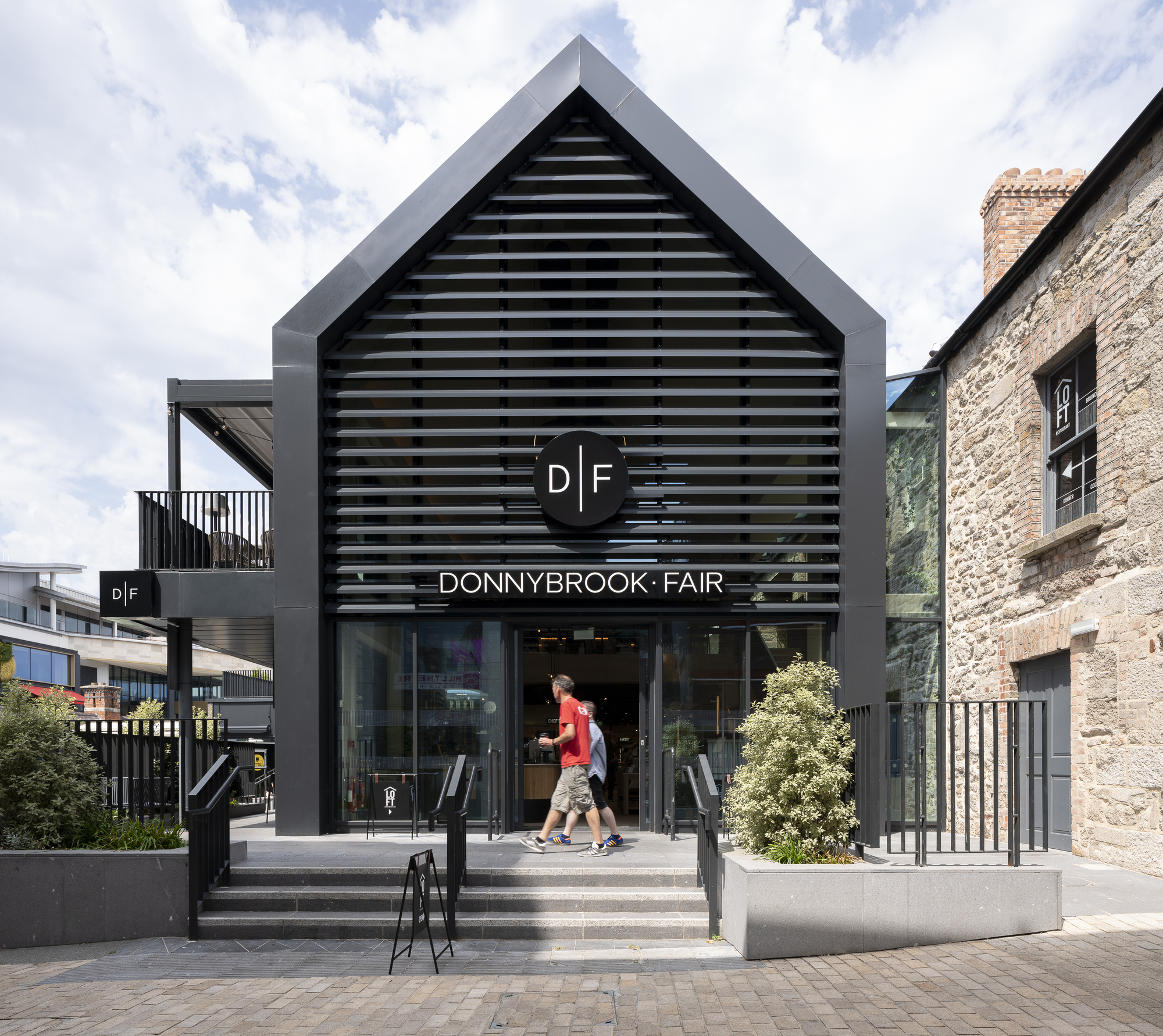 Gable-fronted entrance with horizontal black louvers and pedestrians ascending concrete steps under cloudy sky