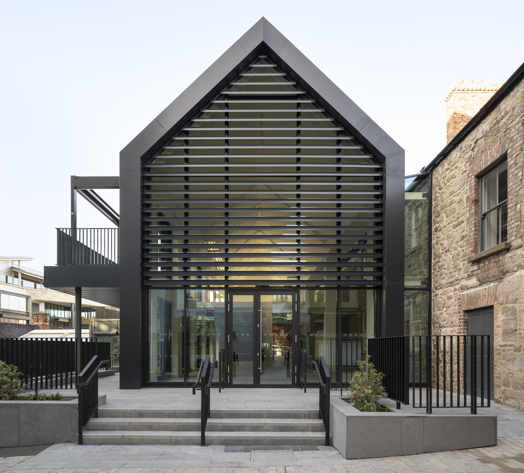 Front elevation of gabled volume with horizontal louvre screen above glazed entrance and stone masonry wing