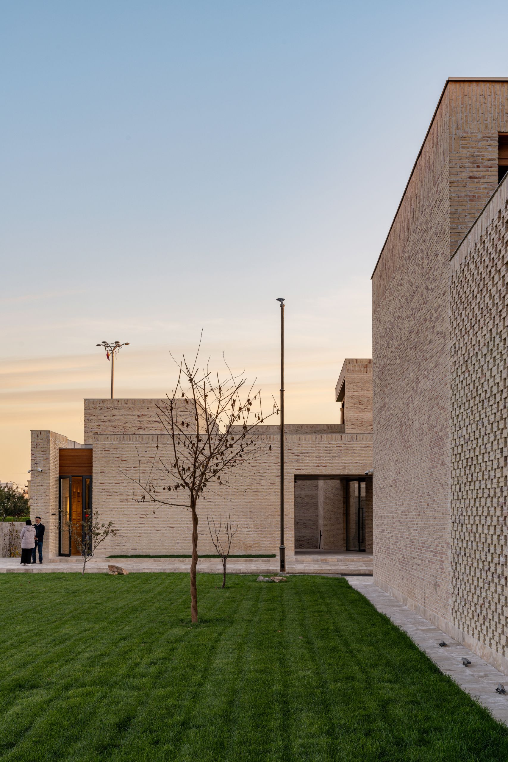 Courtyard view with patterned brick volumes, lawn, and young trees at dusk