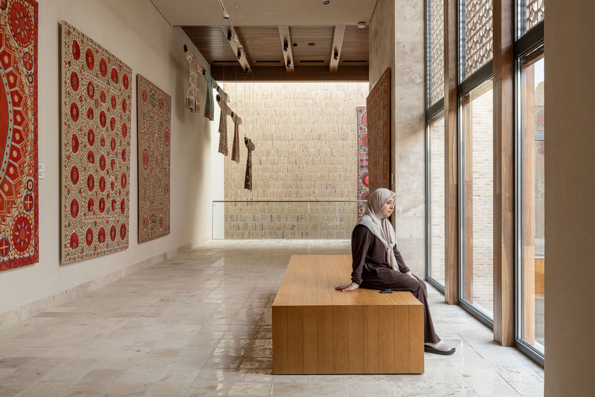 Gallery space with timber bench, stone floor, red textile wall hangings and a seated visitor by the window